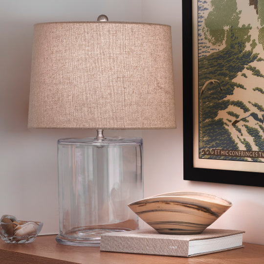 Simon Pearce handblown cylindrical glass table lamp with a linen shade on a wood shelf, styled with a decorative glass bowl and framed art