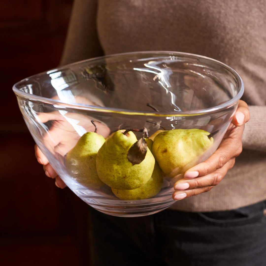 Three Simon Pearce handblown glass bowls in graduated sizes filled with fresh fruit on a green linen tablecloth near a sunlit window