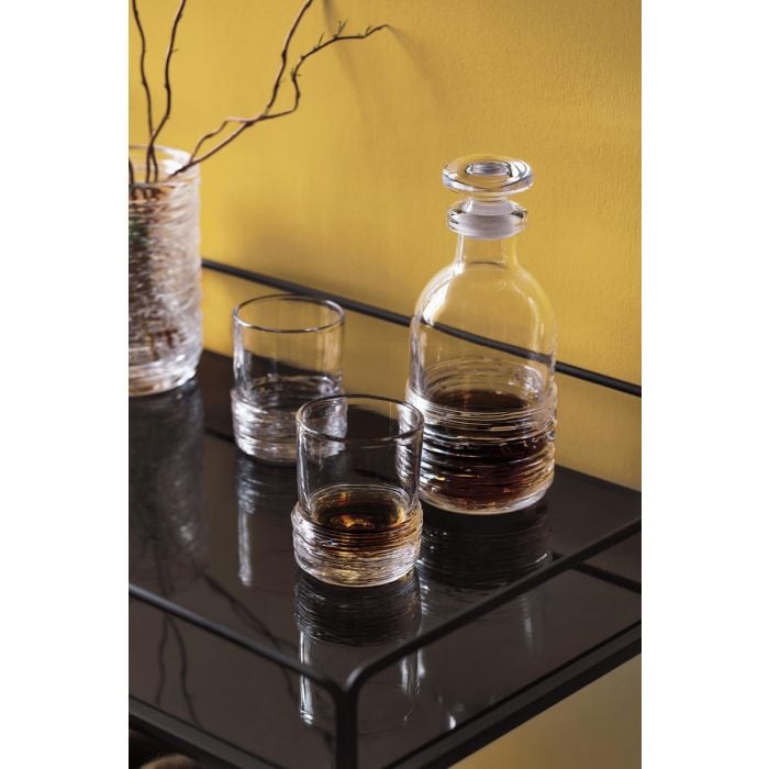 Glass decanter with stopper and two whiskey glasses filled with amber liquid on black table against yellow wall.