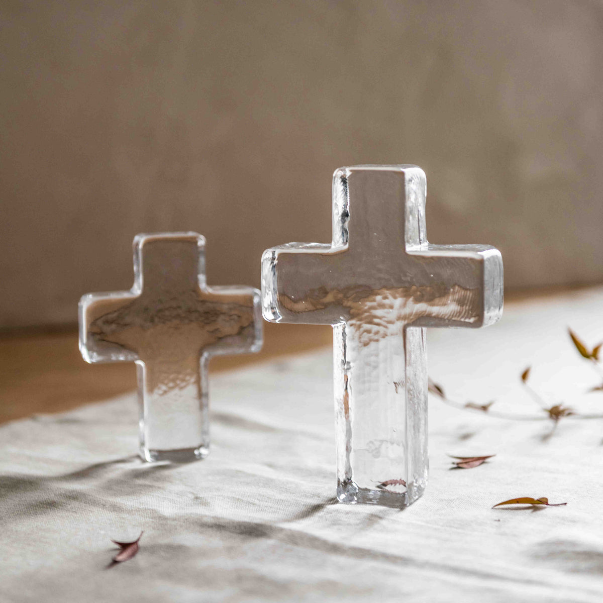 Two clear glass crosses standing on a white surface with dried leaves scattered nearby.