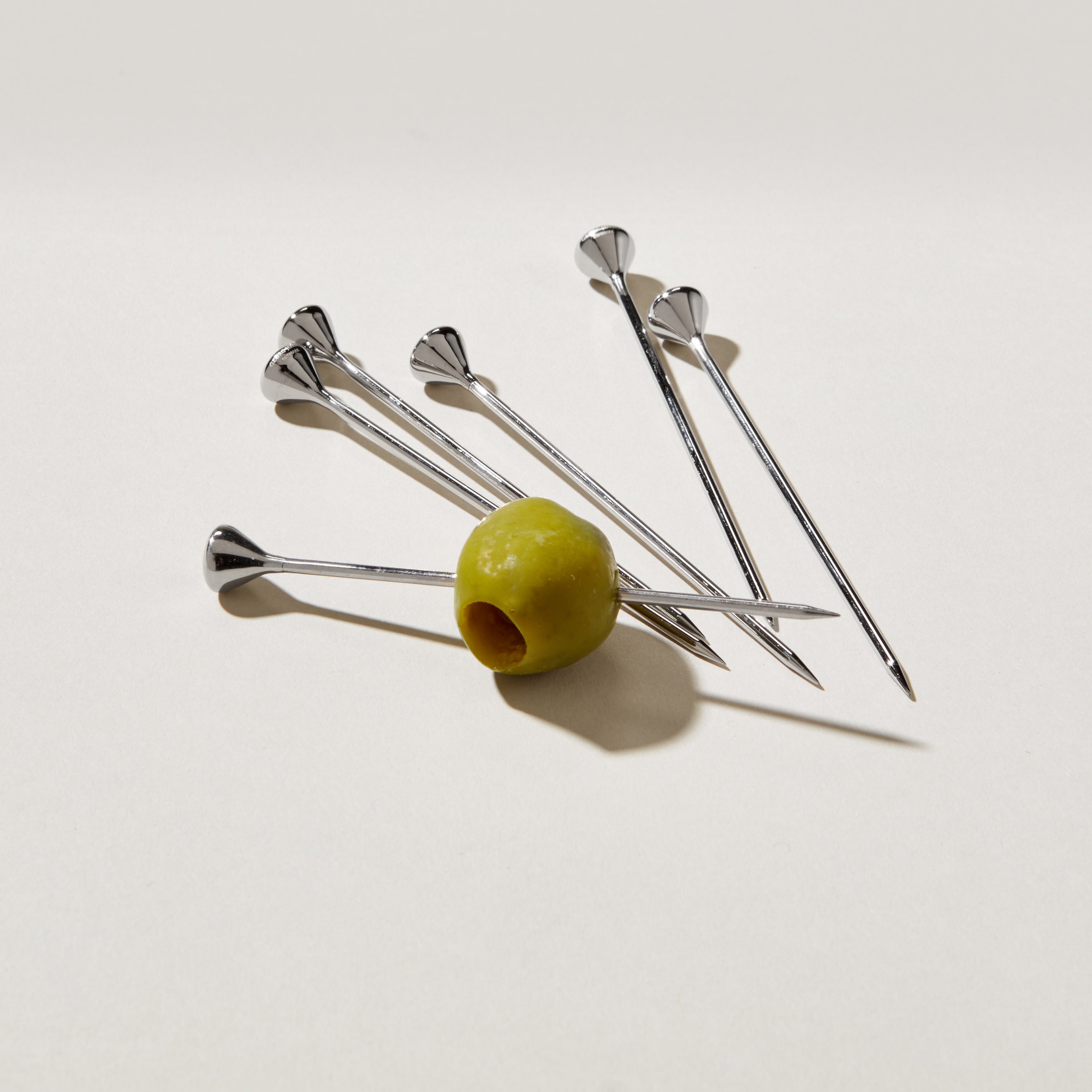 Six stainless steel cocktail picks with decorative fan-shaped tops, one holding a green olive.