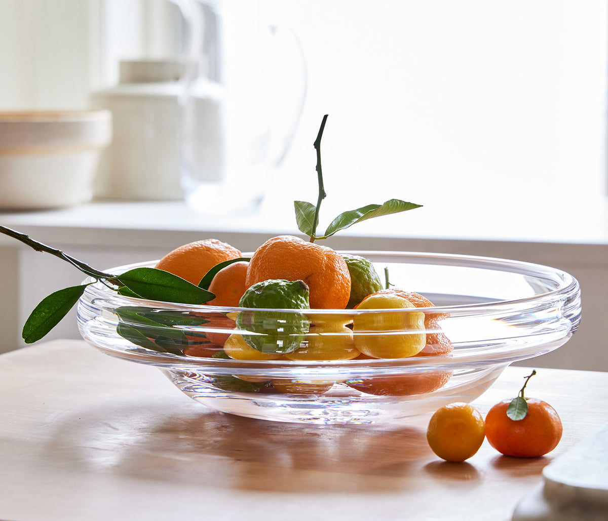 Clear glass platter with wide shallow bowl containing small oranges and green branches.