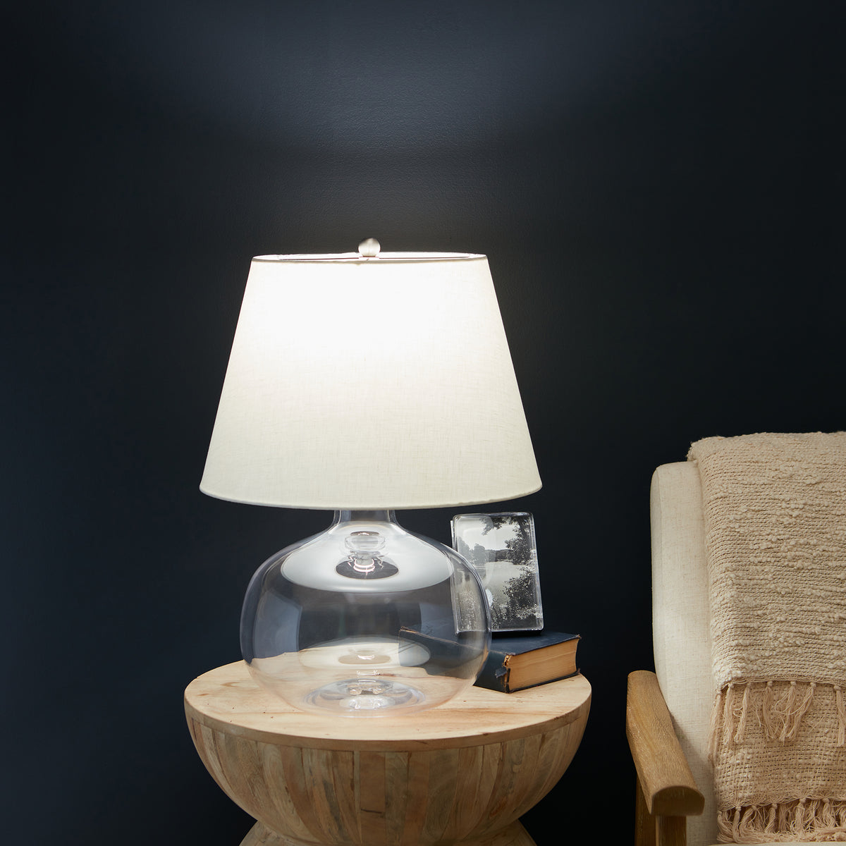 Round table lamp with clear glass base, white fabric shade, and metal finial on wooden surface.