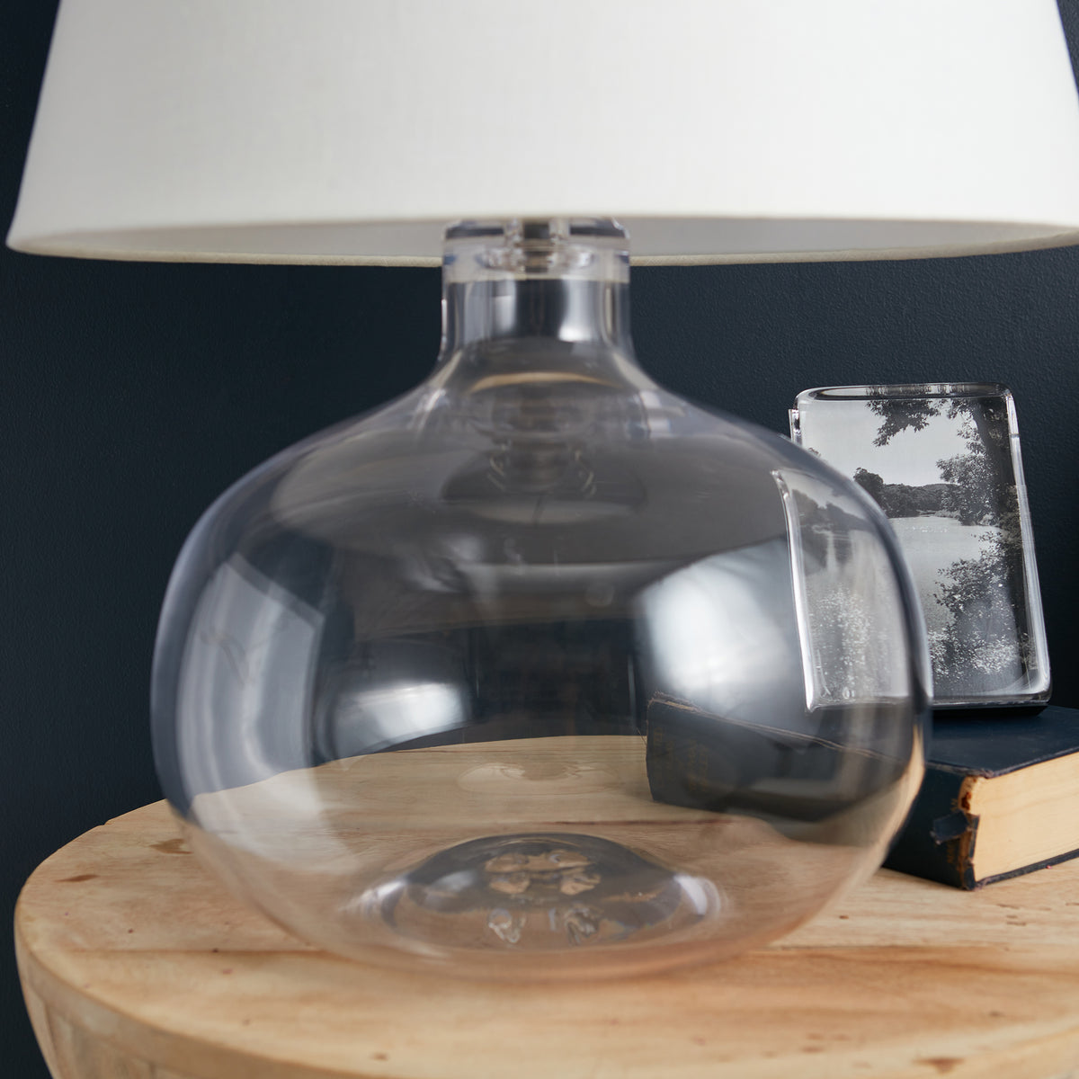 Clear glass round base lamp with white cylindrical shade and metal fixture on wooden table.