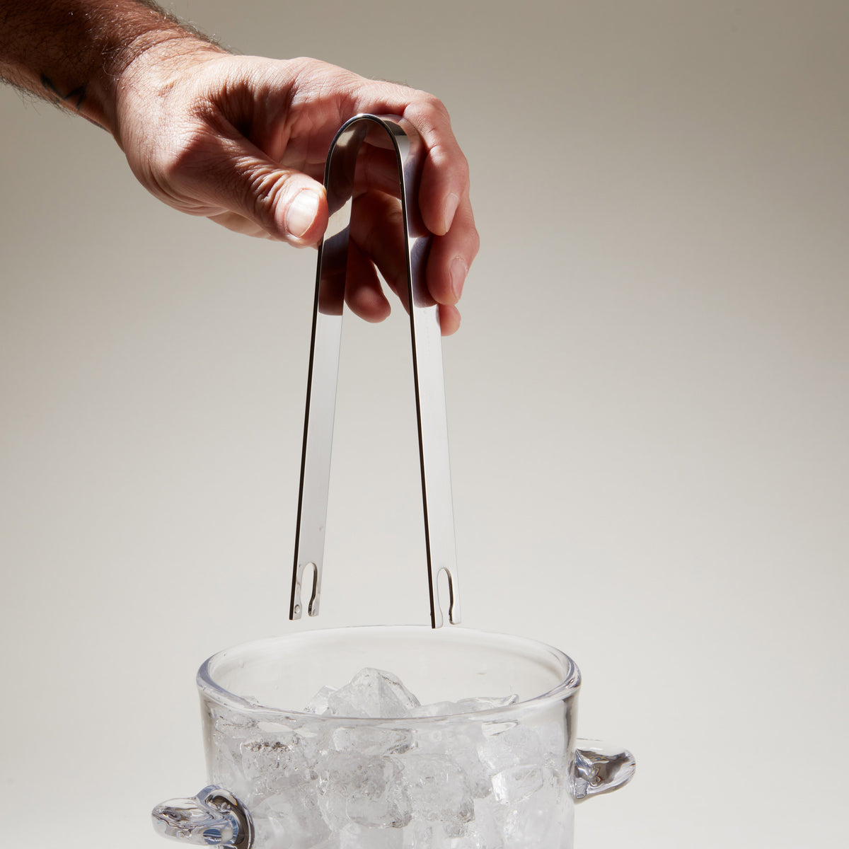 Hand holding stainless steel ice tongs with curved handles over a glass filled with ice cubes.