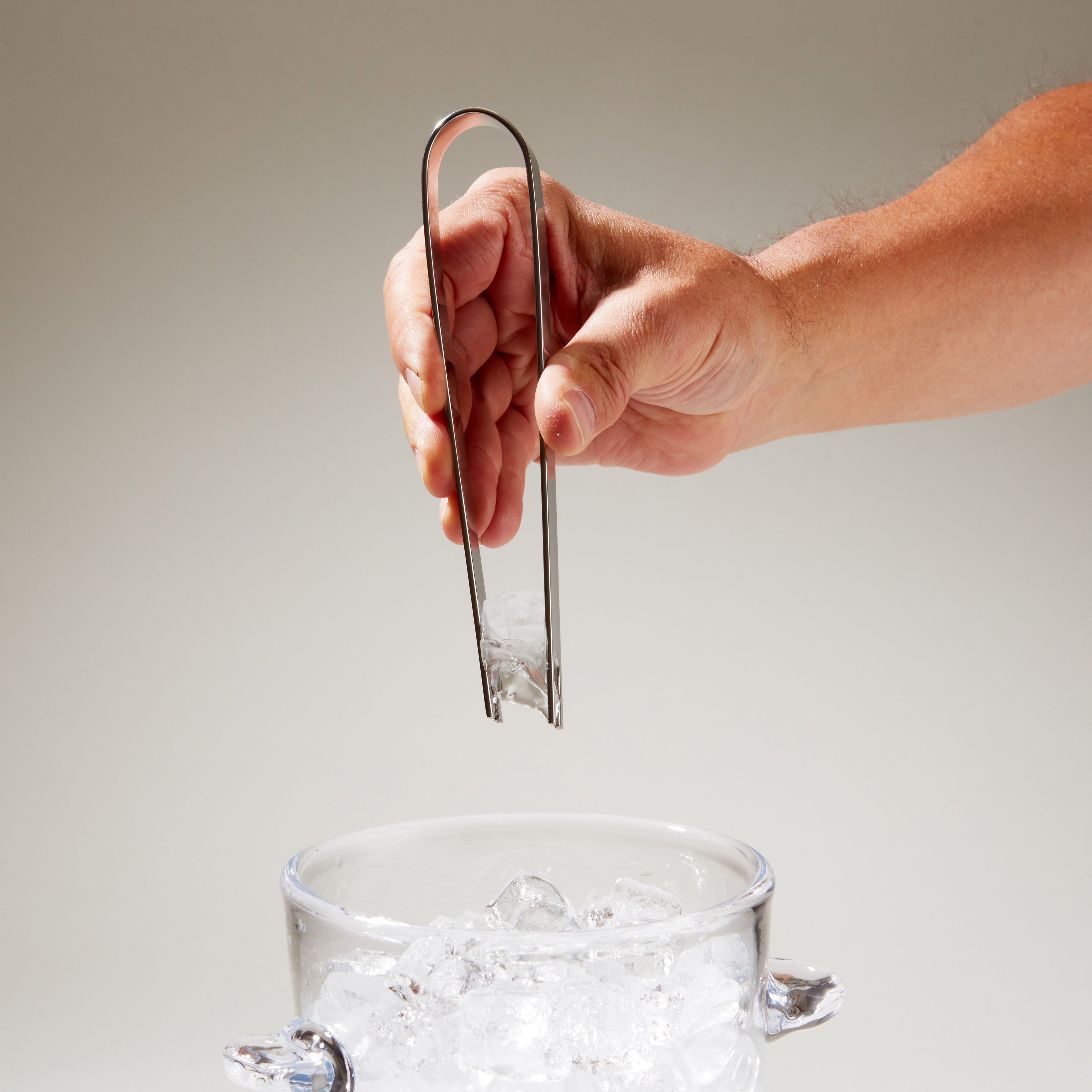 Stainless steel ice tongs held in hand above a glass of ice cubes.