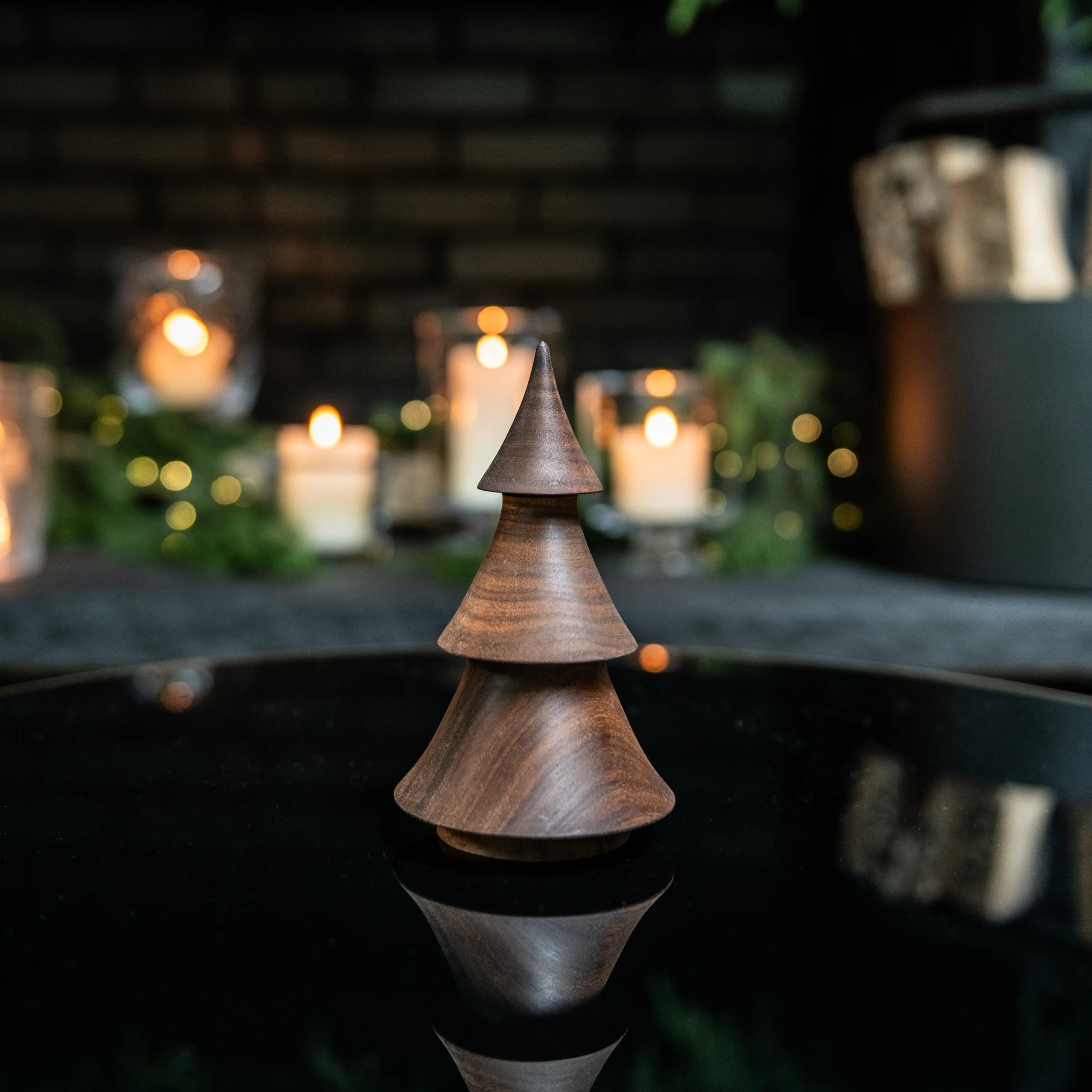 Stacked walnut wood evergreen tree decoration with pointed top, displayed on dark surface with blurred candles and greenery i
