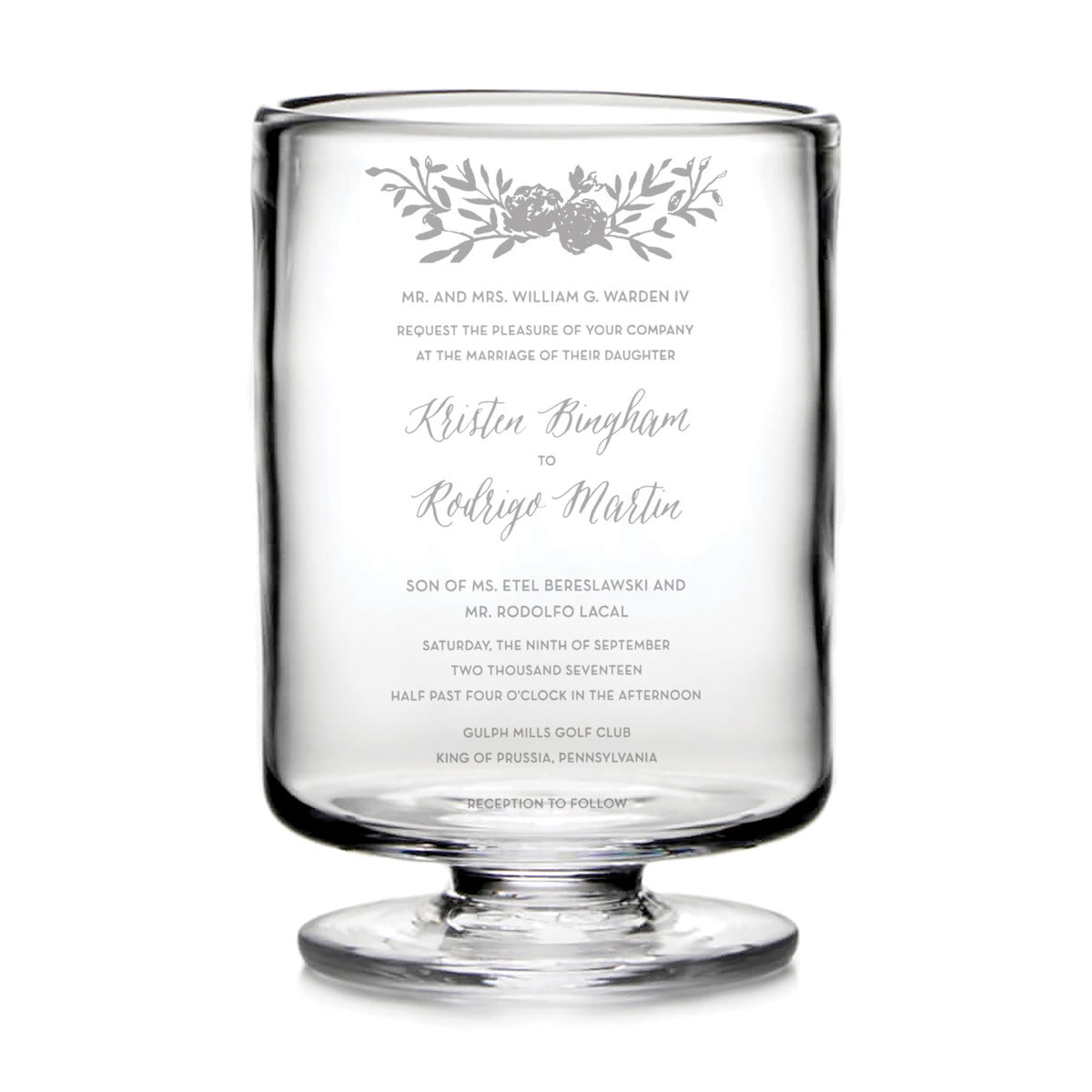 Clear hurricane glass with engraved wedding invitation text and floral design at top.