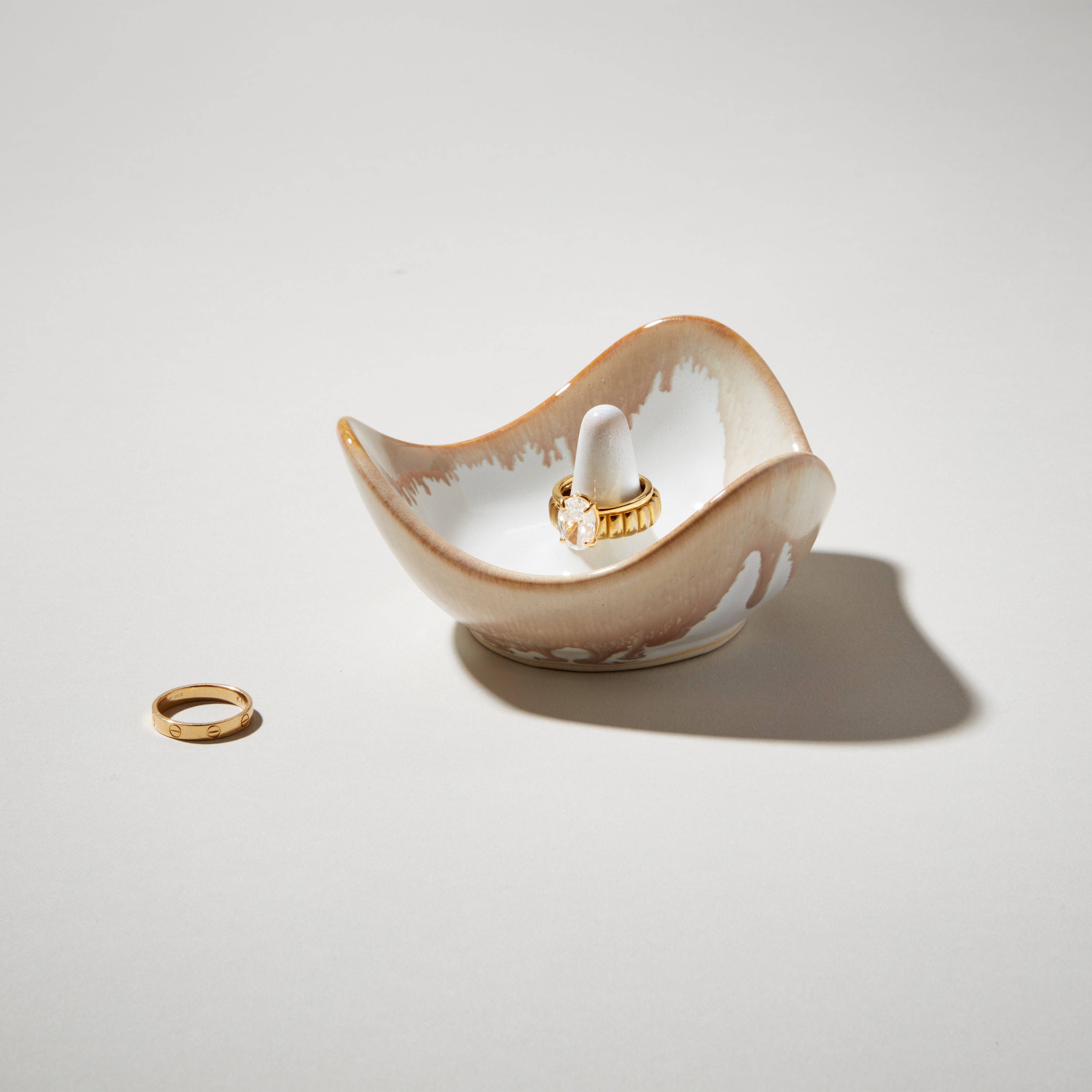 Curved ceramic ring holder in cream and tan with white interior, displaying two gold-toned rings.