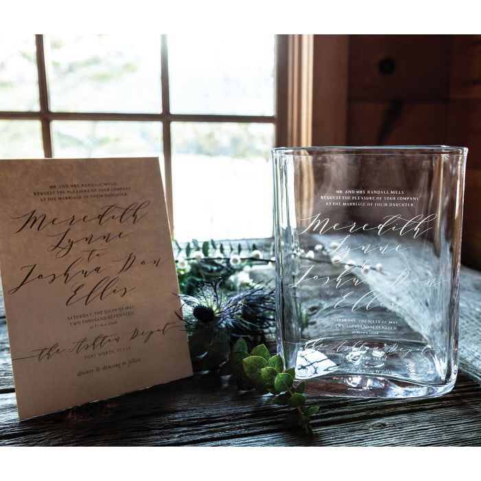 Clear glass vase with engraved wedding invitation text and names, displayed next to tan invitation card.