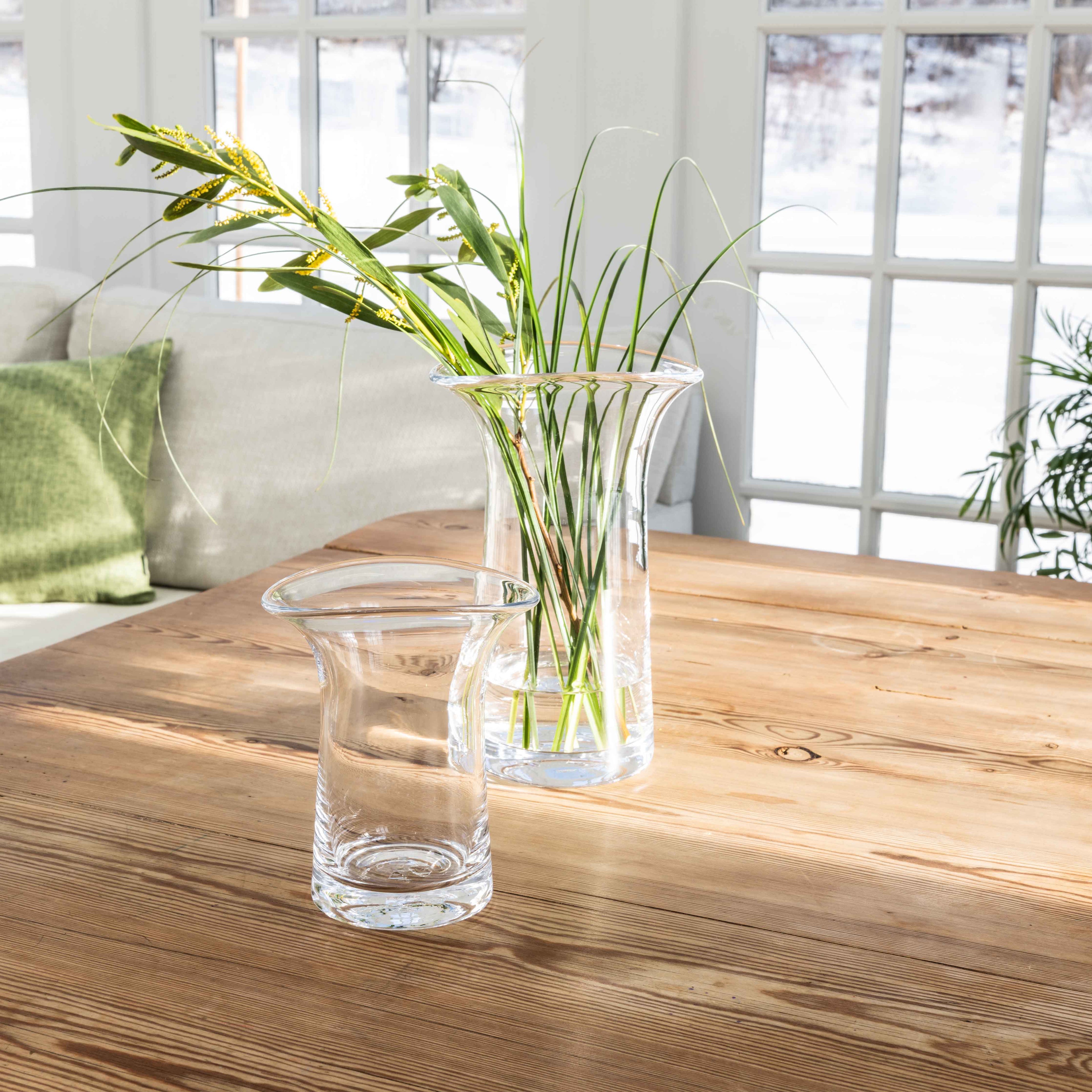 Clear glass vase with straight sides holding green stems and grasses on wooden table.