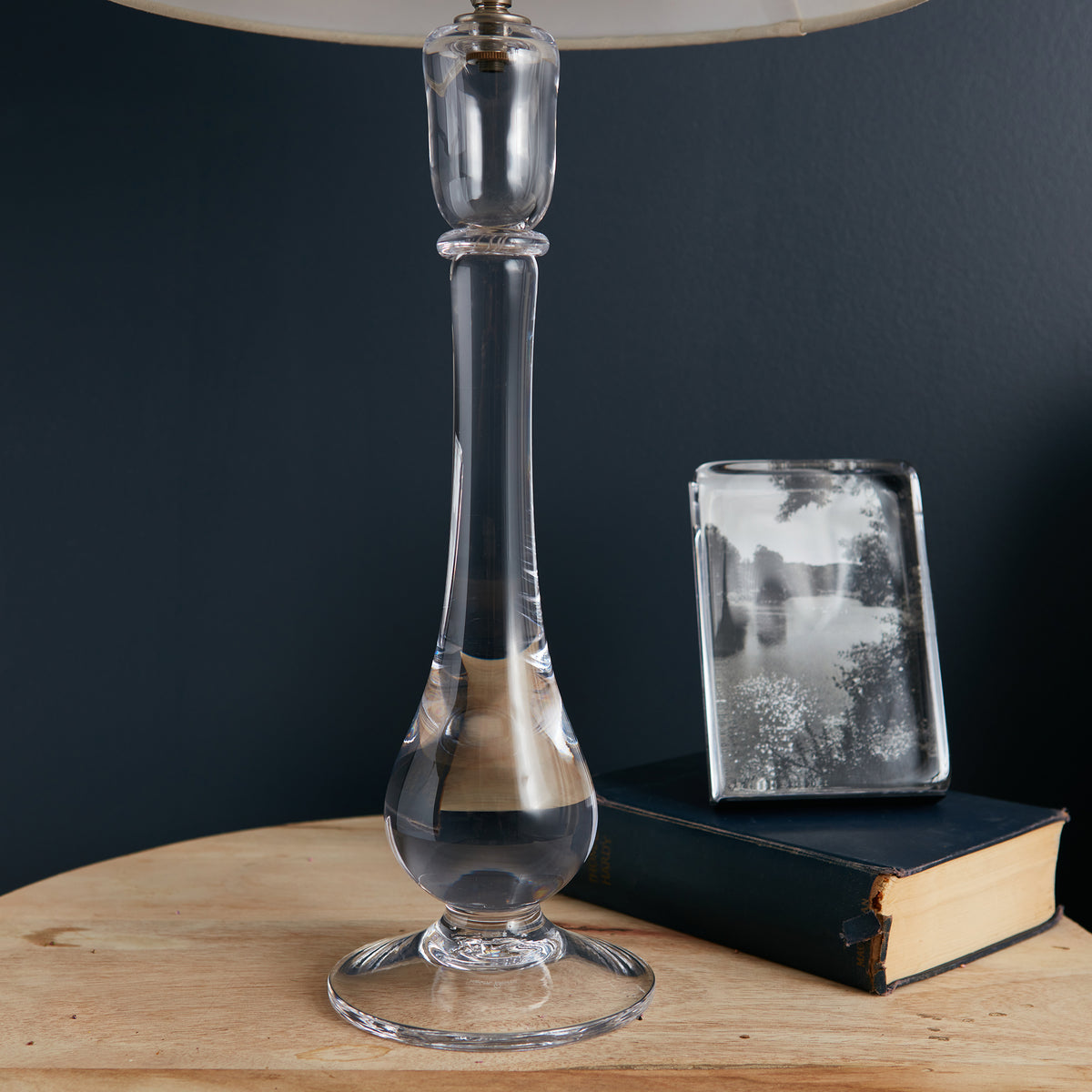 Clear glass table lamp with tapered stem, brass band detail, and cream linen shade.
