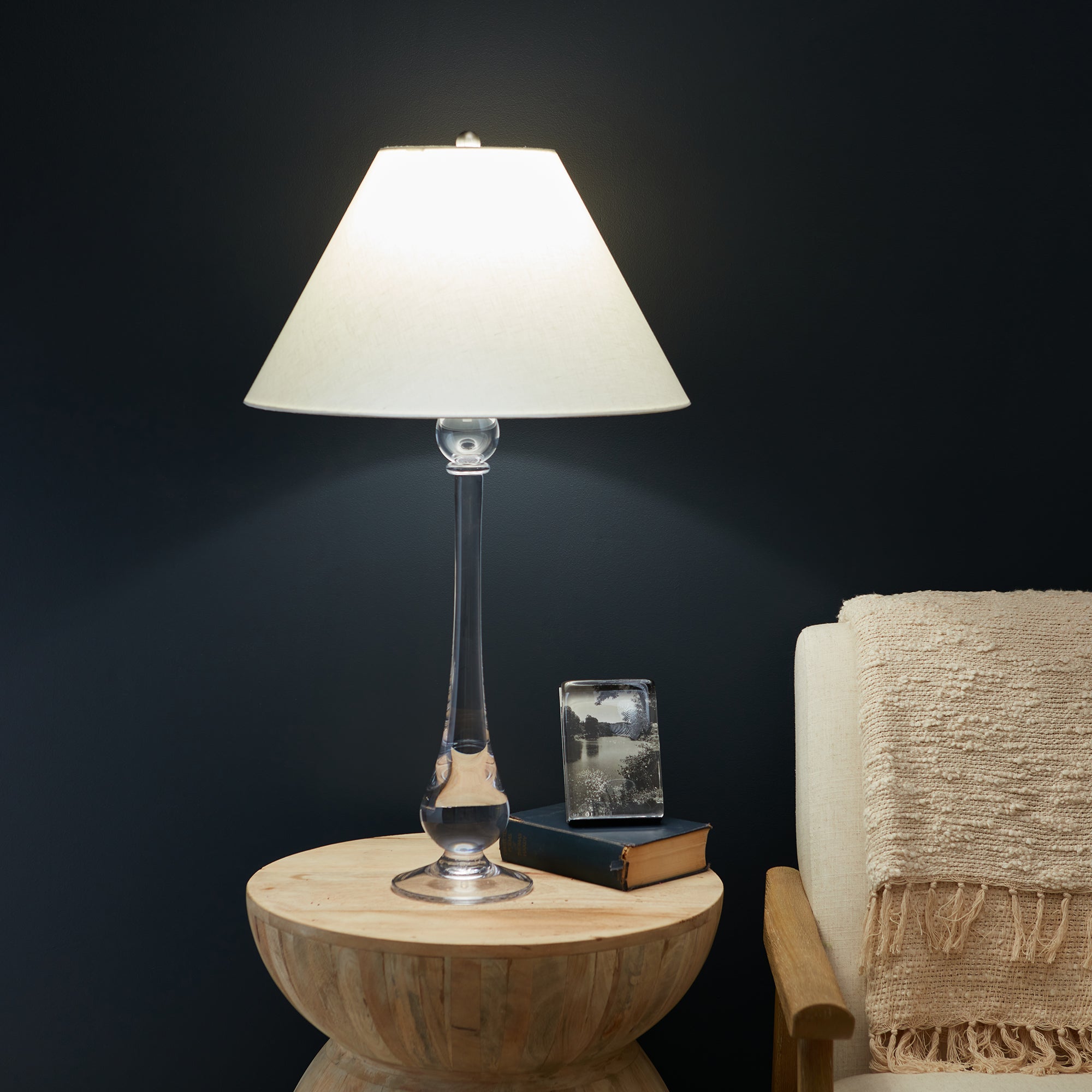 Tall lamp with cream linen trapezoid shade, clear glass and metal stem base, on wooden side table.