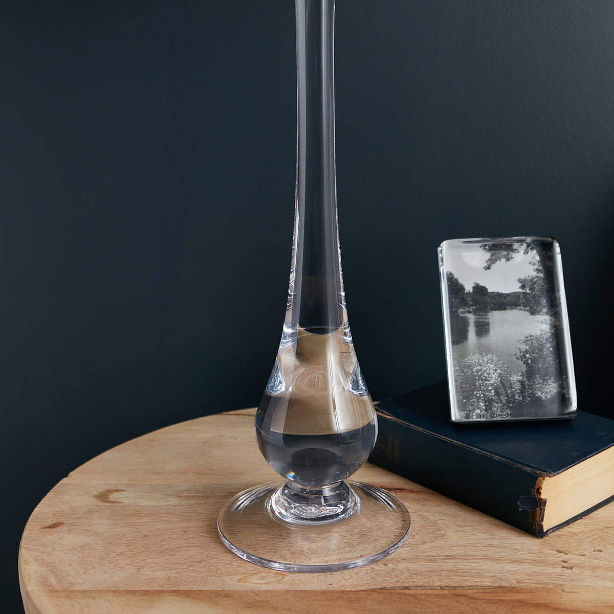Tall glass lamp with teardrop-shaped shade and wooden base, displayed on round wooden table next to books.