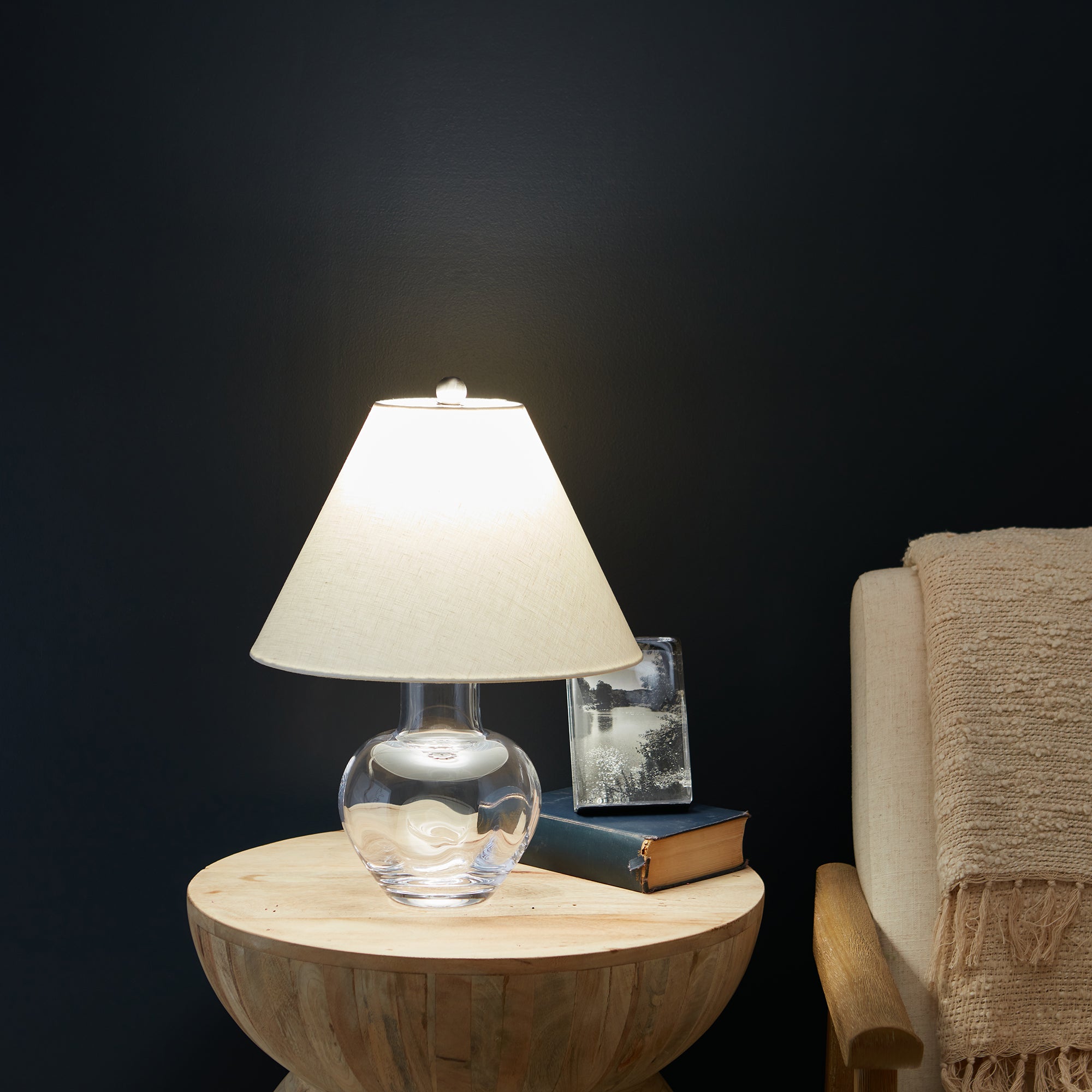 Small table lamp with clear glass bulbous base, cream linen shade, and finial, displayed on wooden round table.