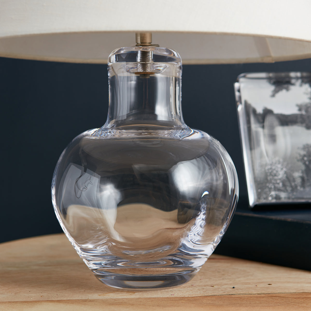 Clear glass bulbous lamp base with brass fixture and cream-colored linen shade on dark background.