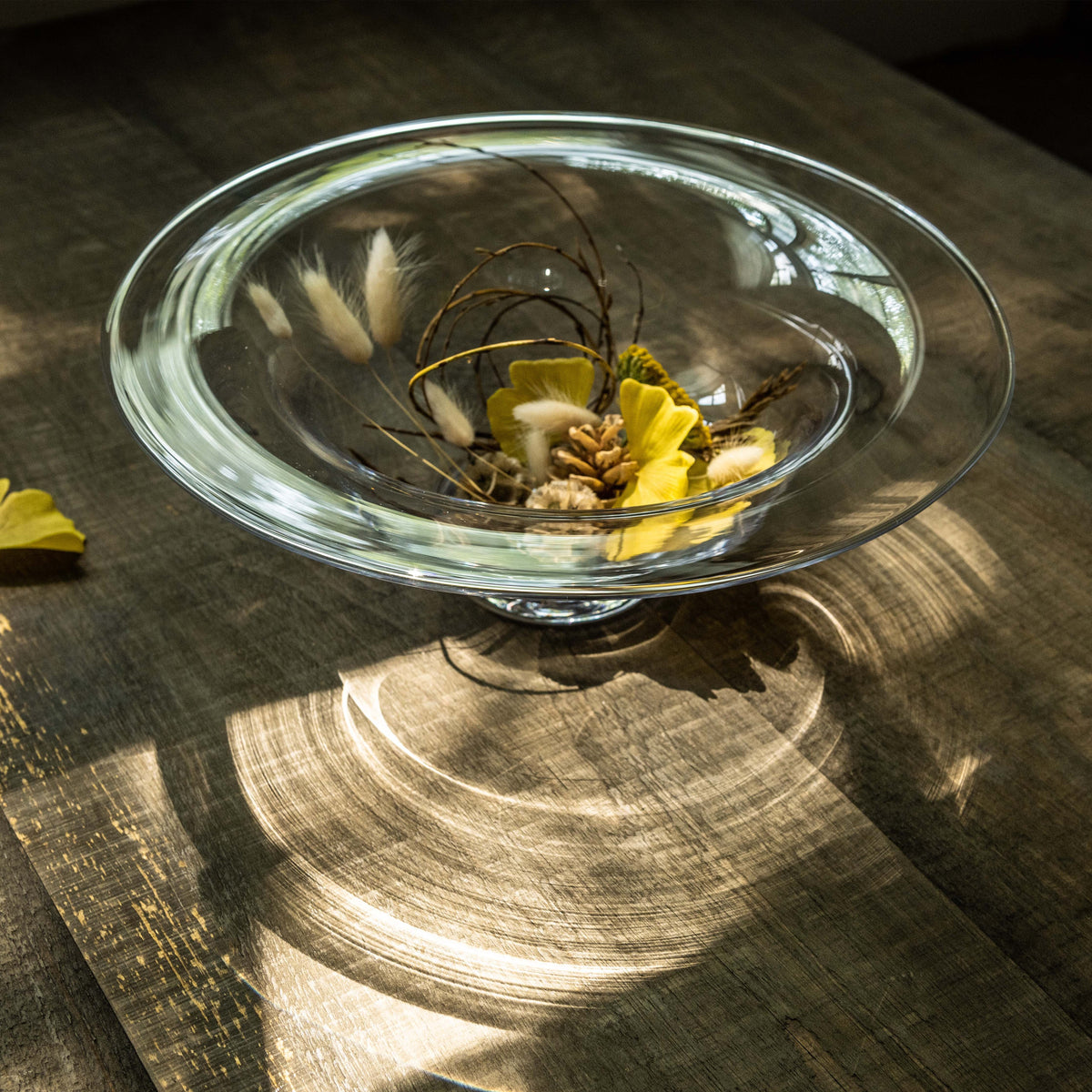 Clear glass pedestal bowl filled with yellow flowers and dried branches on wooden surface.