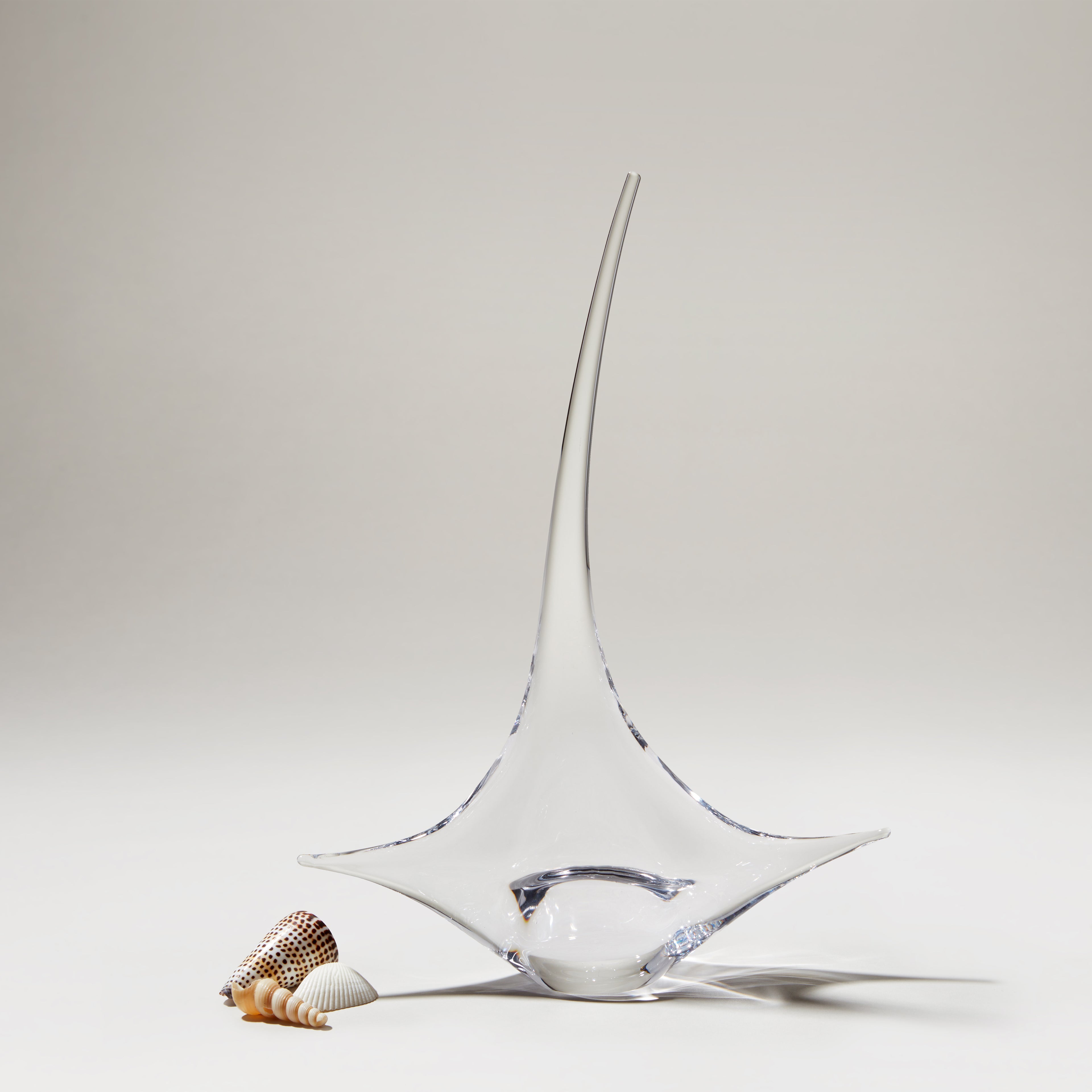Clear glass sailboat sculpture with pointed mast and curved hull base, accompanied by small seashells.