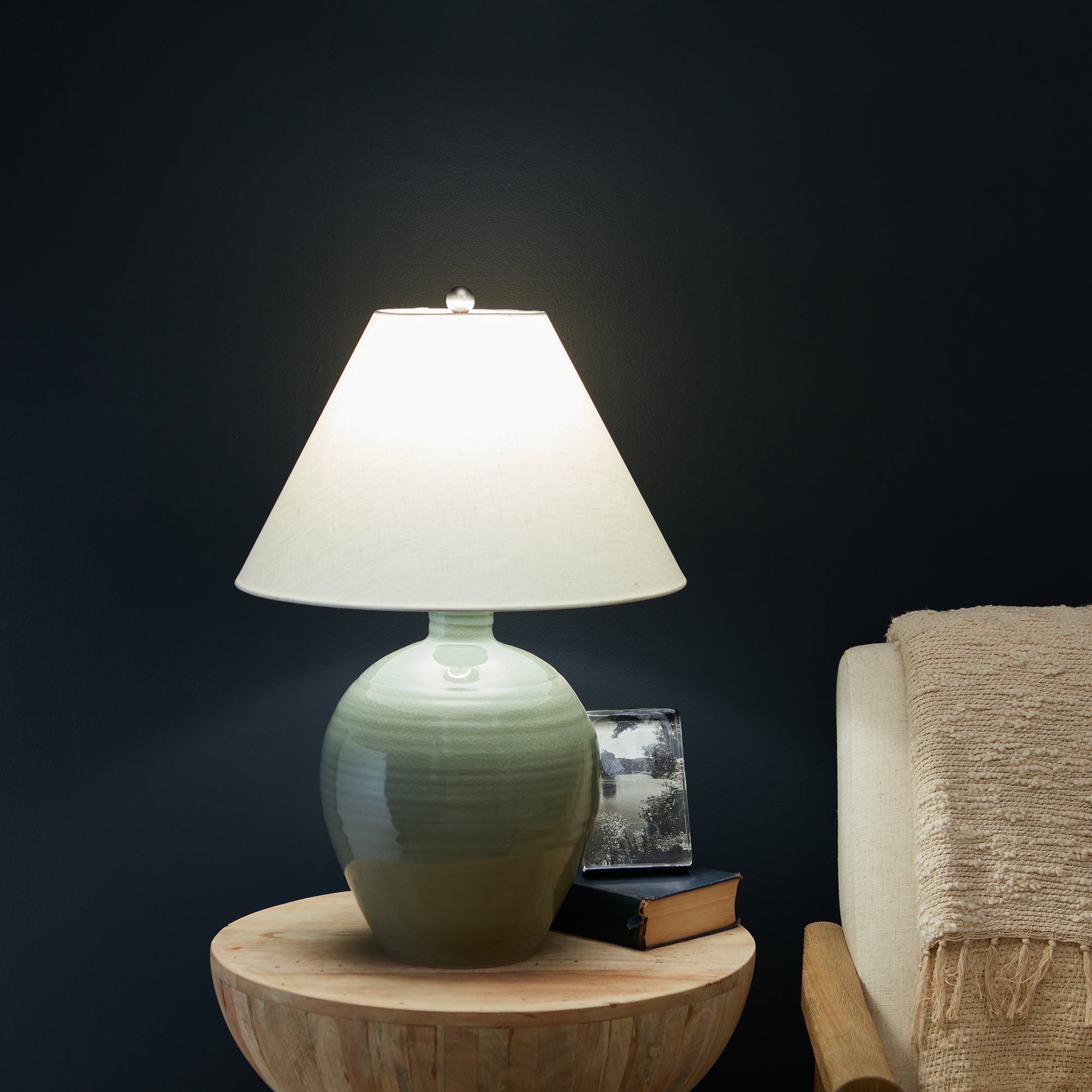 Celadon green ceramic table lamp with white linen shade on wooden base, illuminated against dark background.