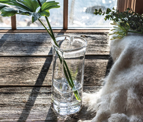Clear glass vase with heart-shaped top holding a green stem, displayed on wooden surface by window.