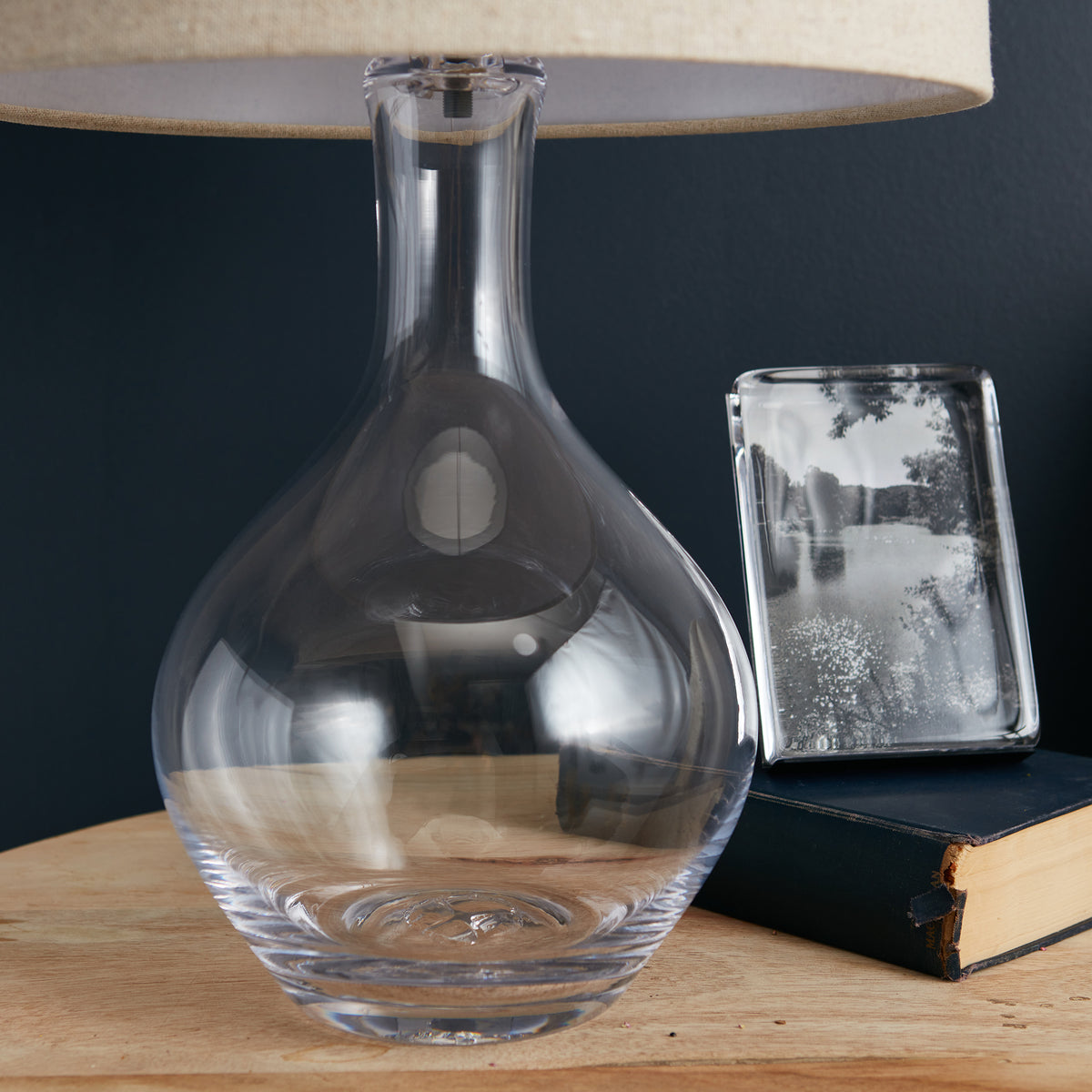 Clear glass teardrop-shaped lamp base with fabric shade, displayed on wooden surface with decorative items.