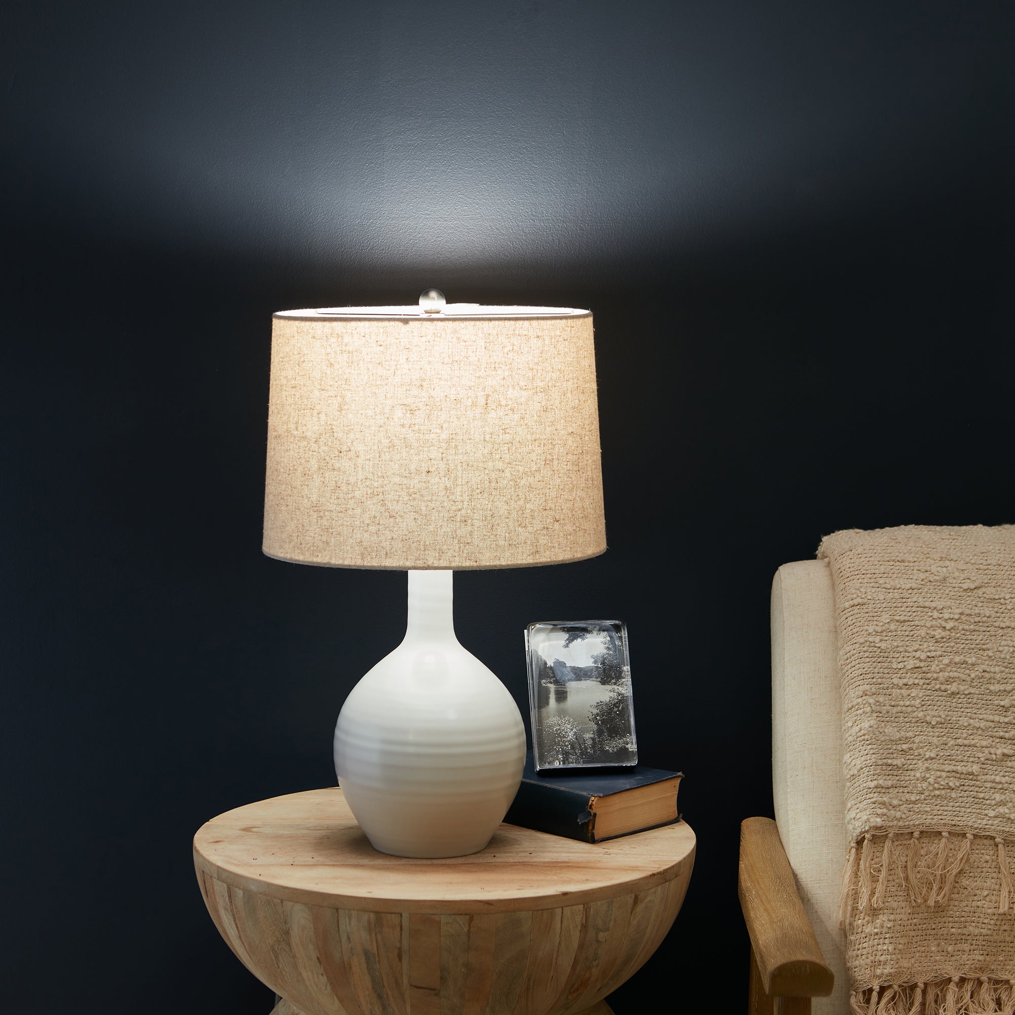 Small white pottery table lamp with rounded base, cylindrical linen shade, and finial top.