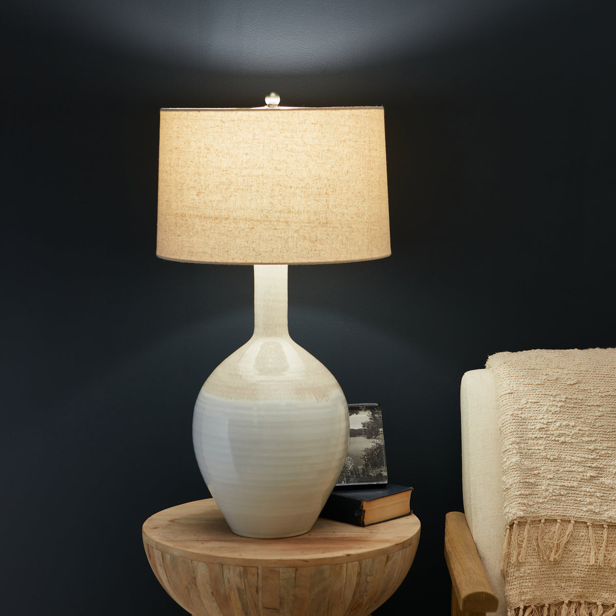 Ceramic table lamp with cream reactive glaze base, narrow neck, and tan linen cylindrical shade on wooden side table.