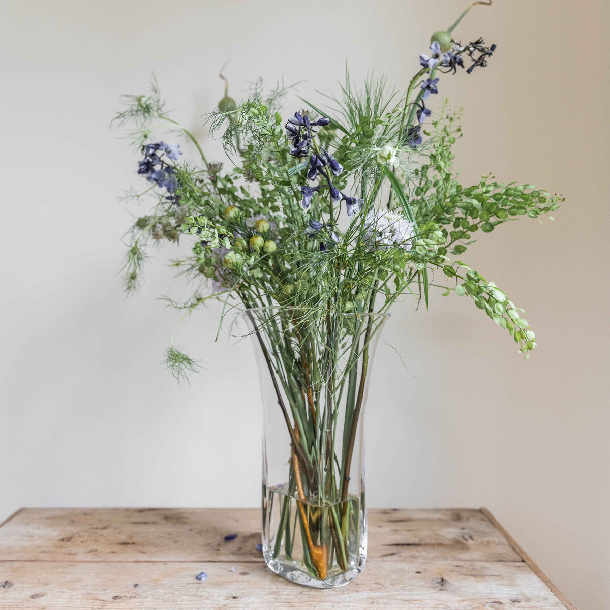 Mixed green foliage and purple flowers arranged in a clear glass vase on a wooden surface.