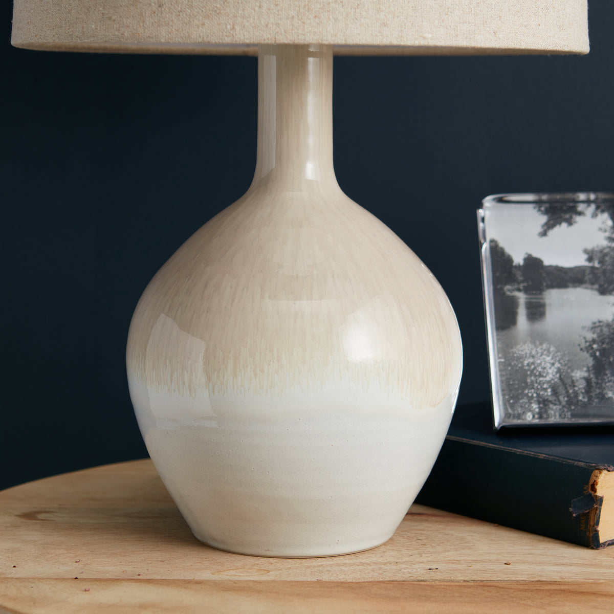 Table lamp with cream reactive glaze ceramic base, narrow neck, and beige linen shade on dark background.