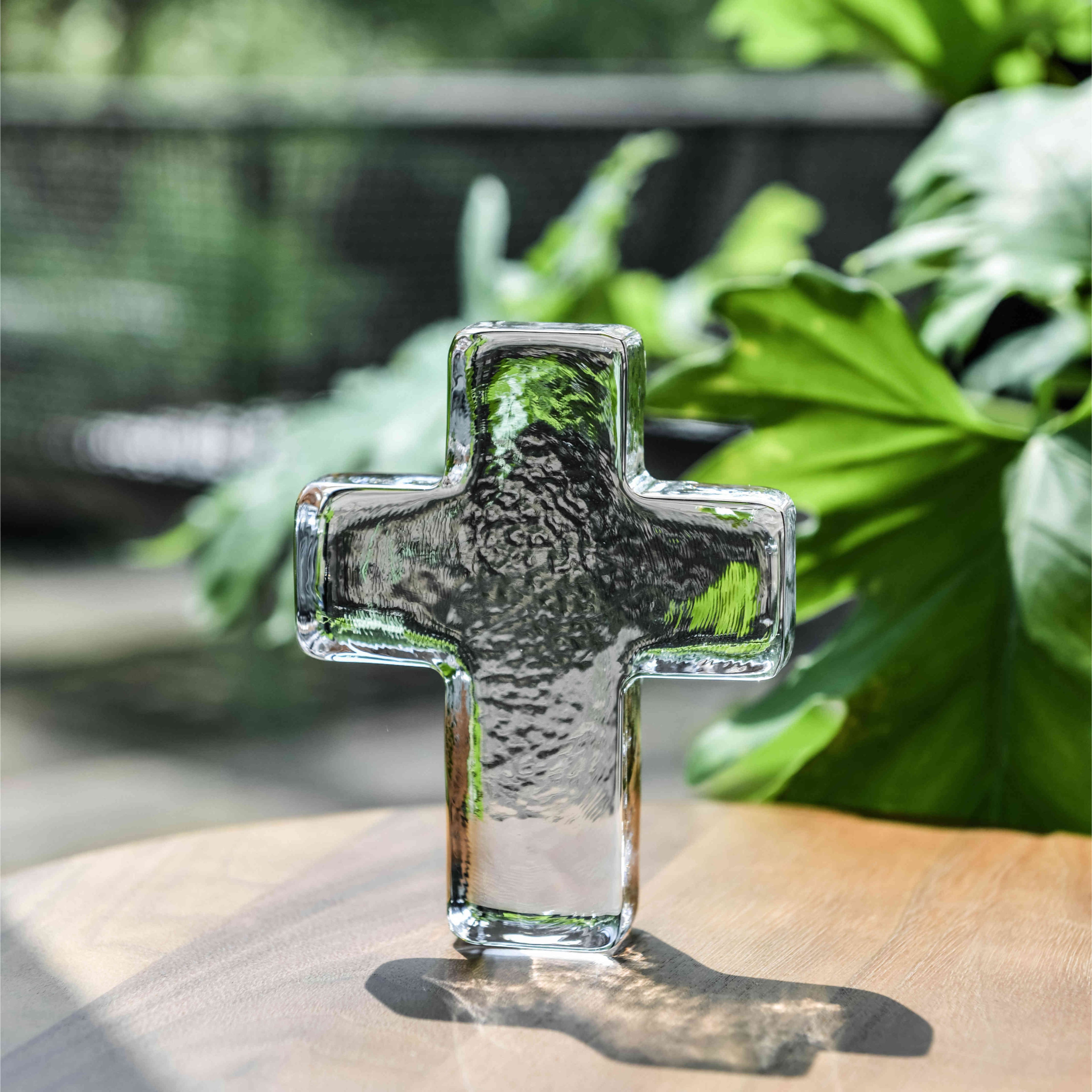 Clear glass cross with textured surface, displayed on wooden surface with green foliage in background.