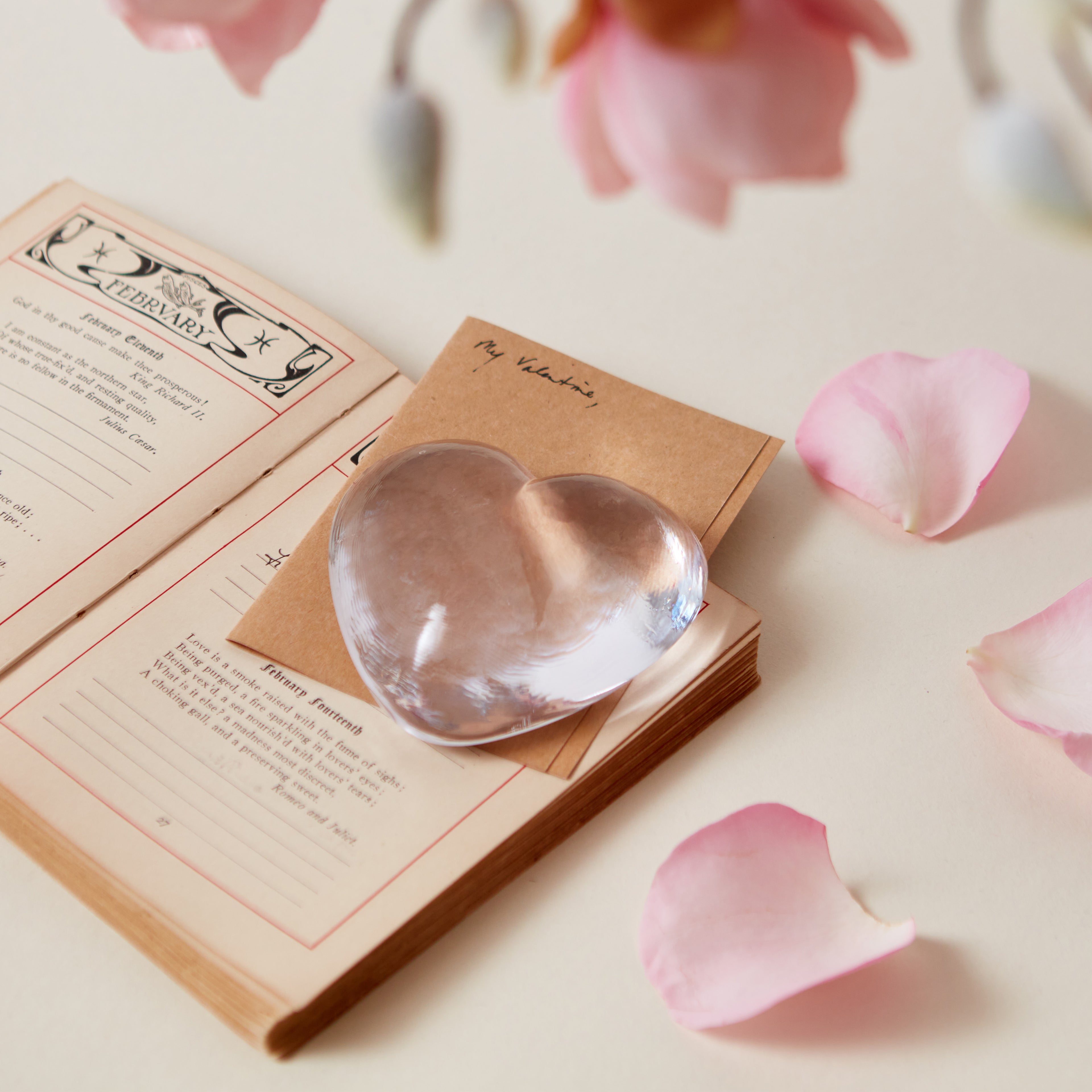 Clear glass heart-shaped paperweight sitting on an open book with pink flower petals scattered nearby.