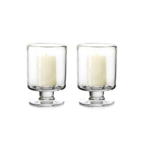 Two clear glass hurricane candle holders with pedestal bases, each containing a cream-colored pillar candle.