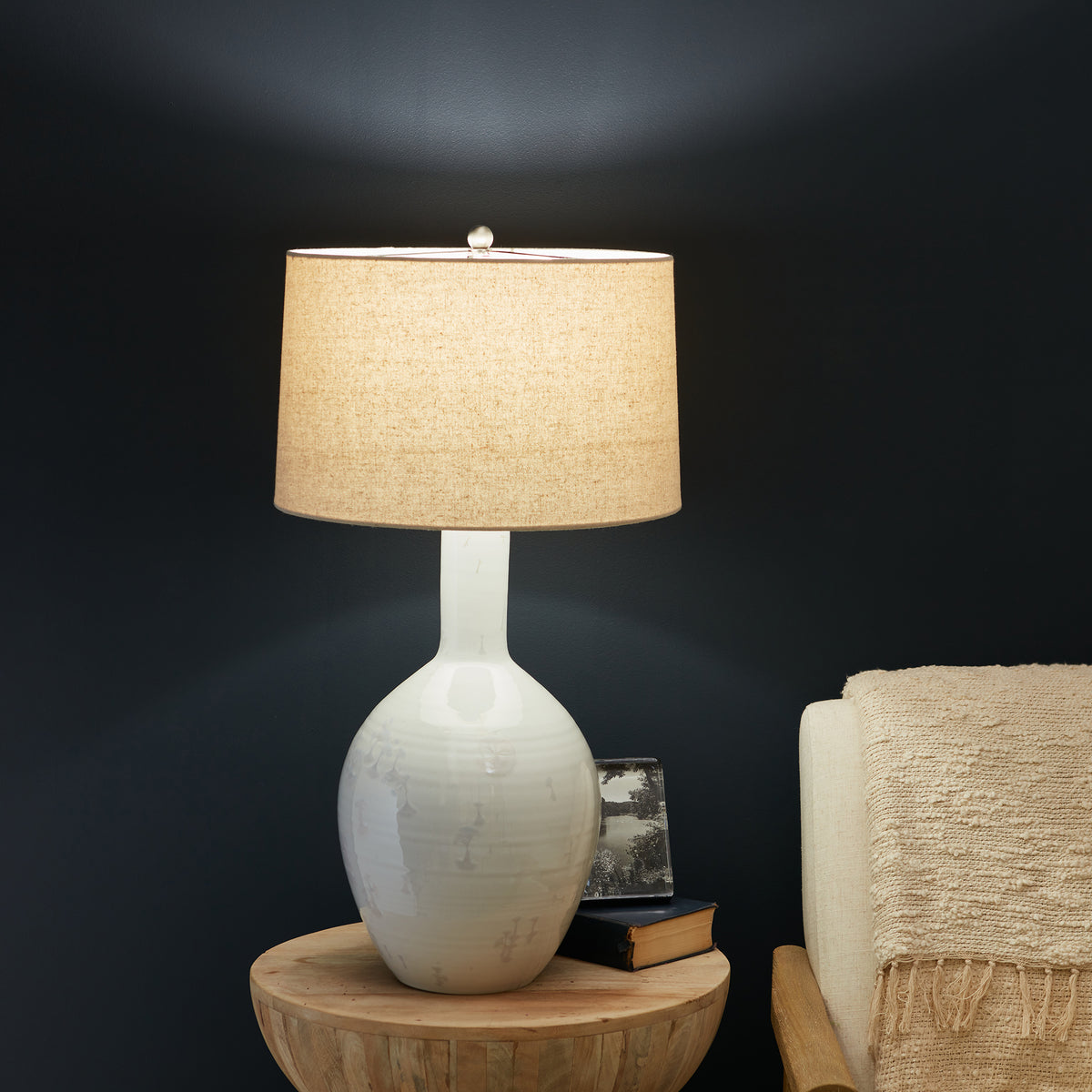 White ceramic lamp base with tan linen drum shade and finial, displayed on wooden table.