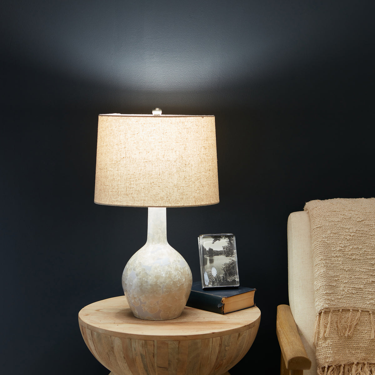 Table lamp with textured white ceramic base, cream linen shade, and silver finial on wooden bowl table.
