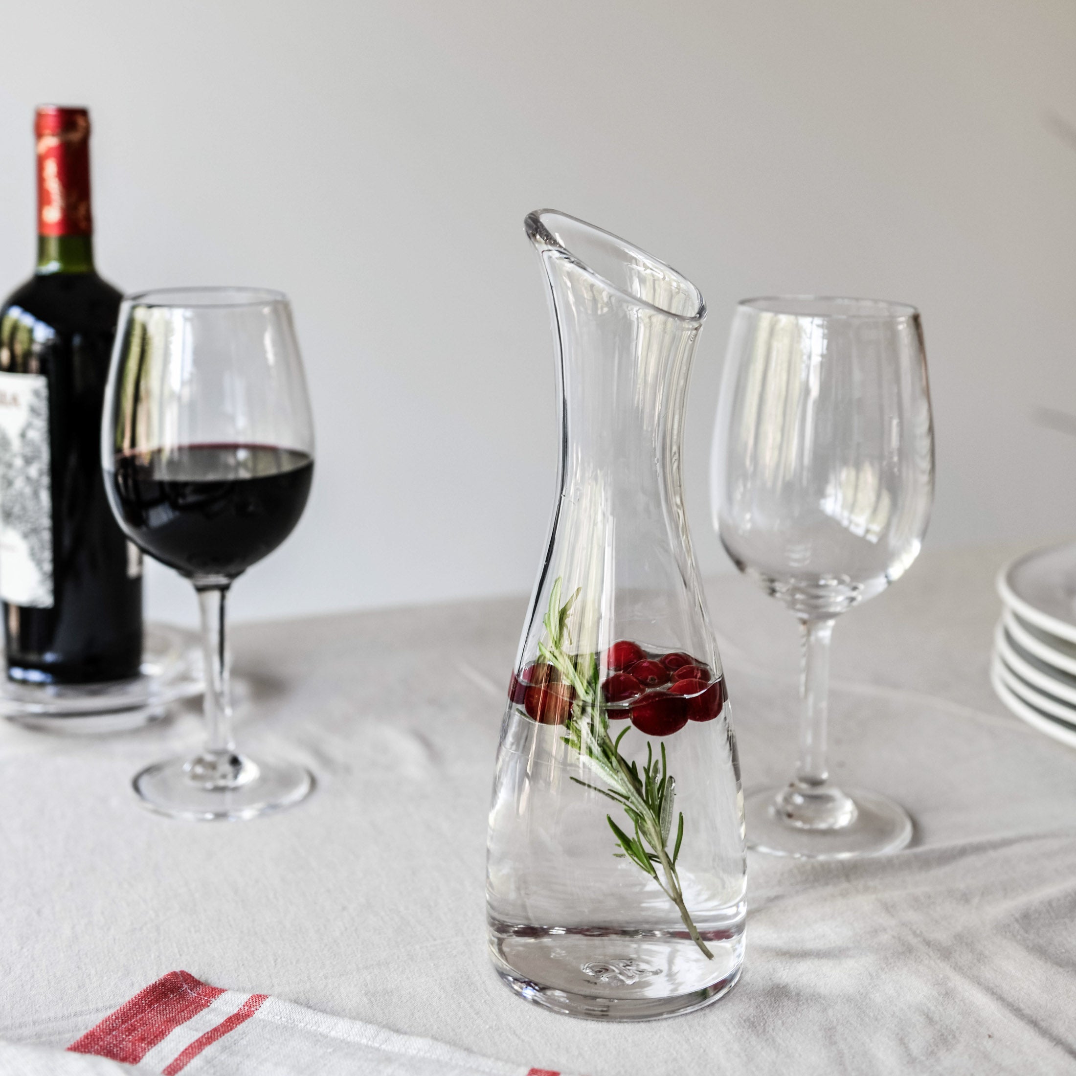 Clear glass carafe with curved body and angled spout, containing water with red berries and green herb sprigs.