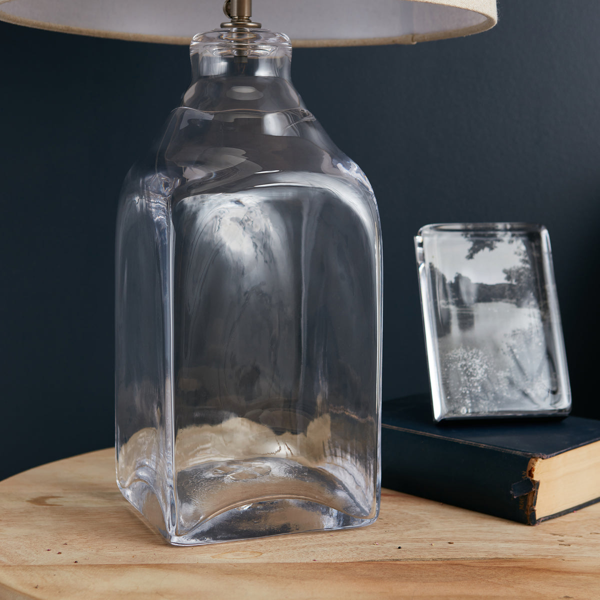 Clear glass bottle lamp base with brass finial and linen shade on wooden surface.