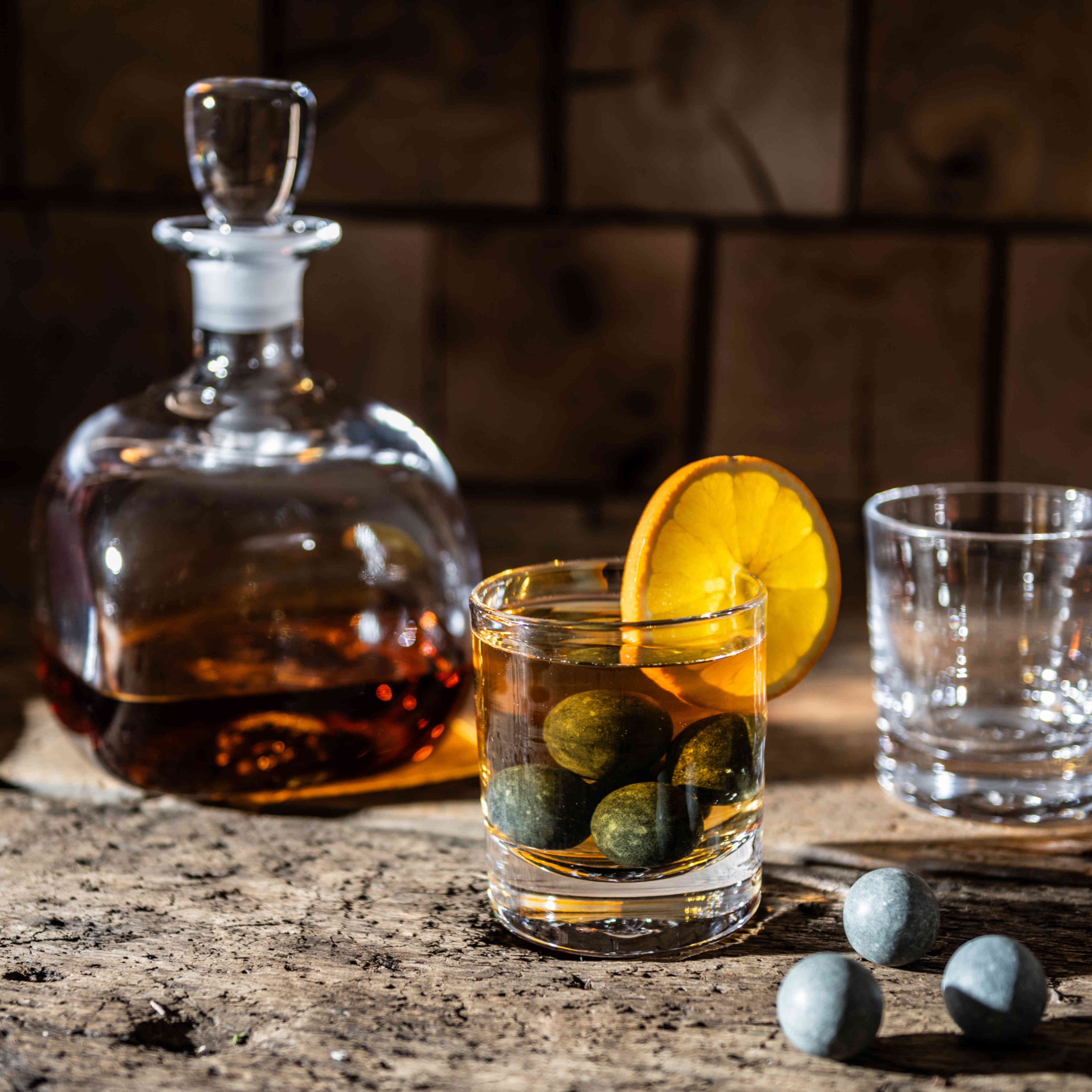 Eight spherical gray stones in a glass of whiskey with lemon slice, whiskey decanter, and empty glasses on stone surface.