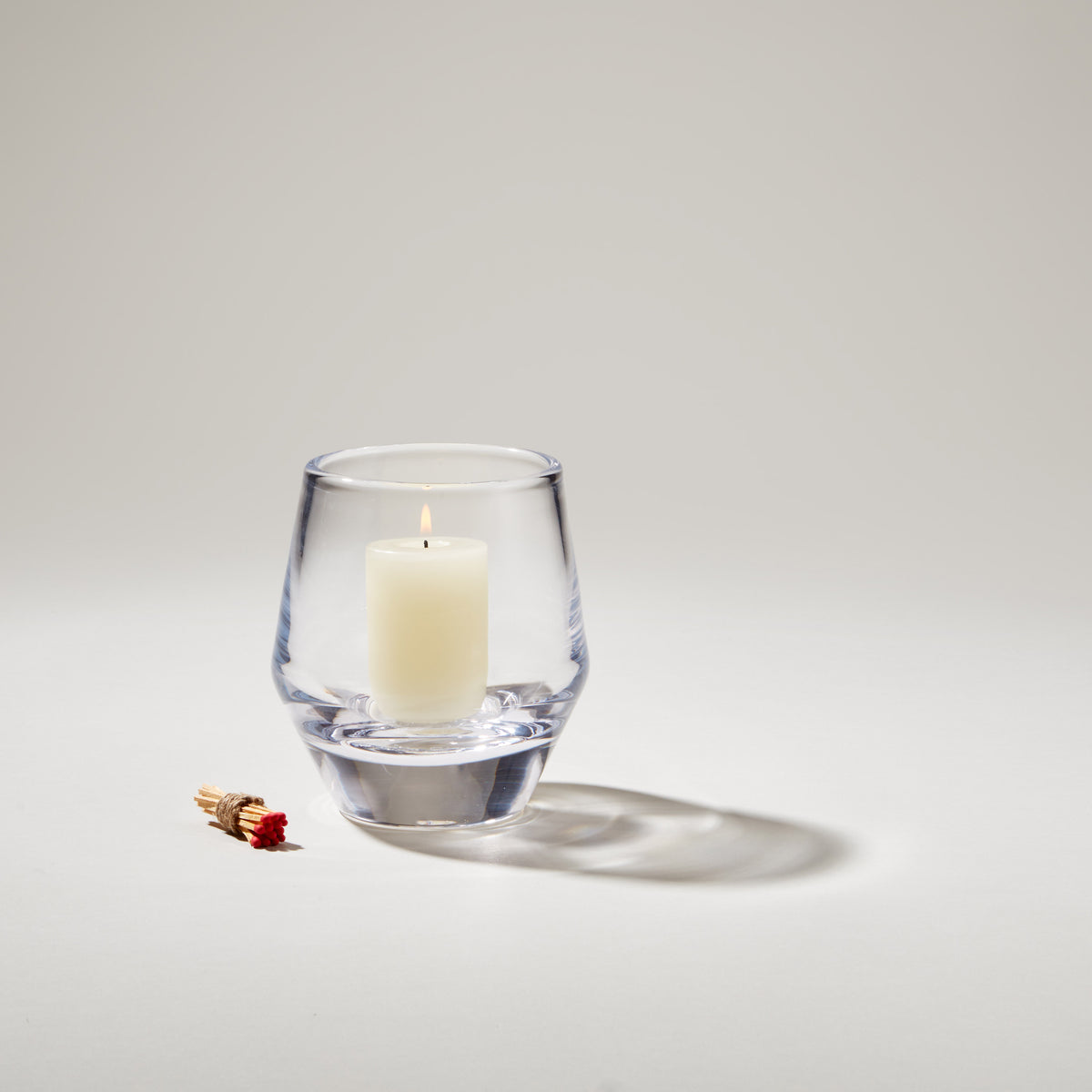 Clear glass votive holder with lit cream-colored candle inside and matches beside it.