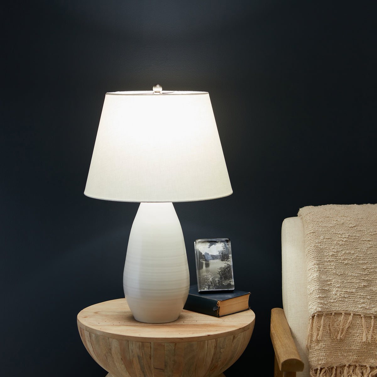 Ceramic table lamp with white textured base and cream linen shade on wooden side table.