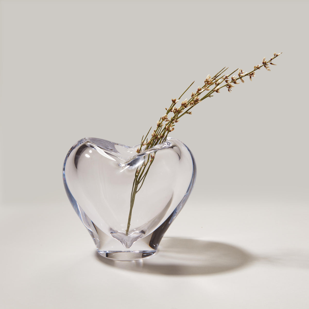 Clear glass heart-shaped vase with dried grasses inside on neutral background.