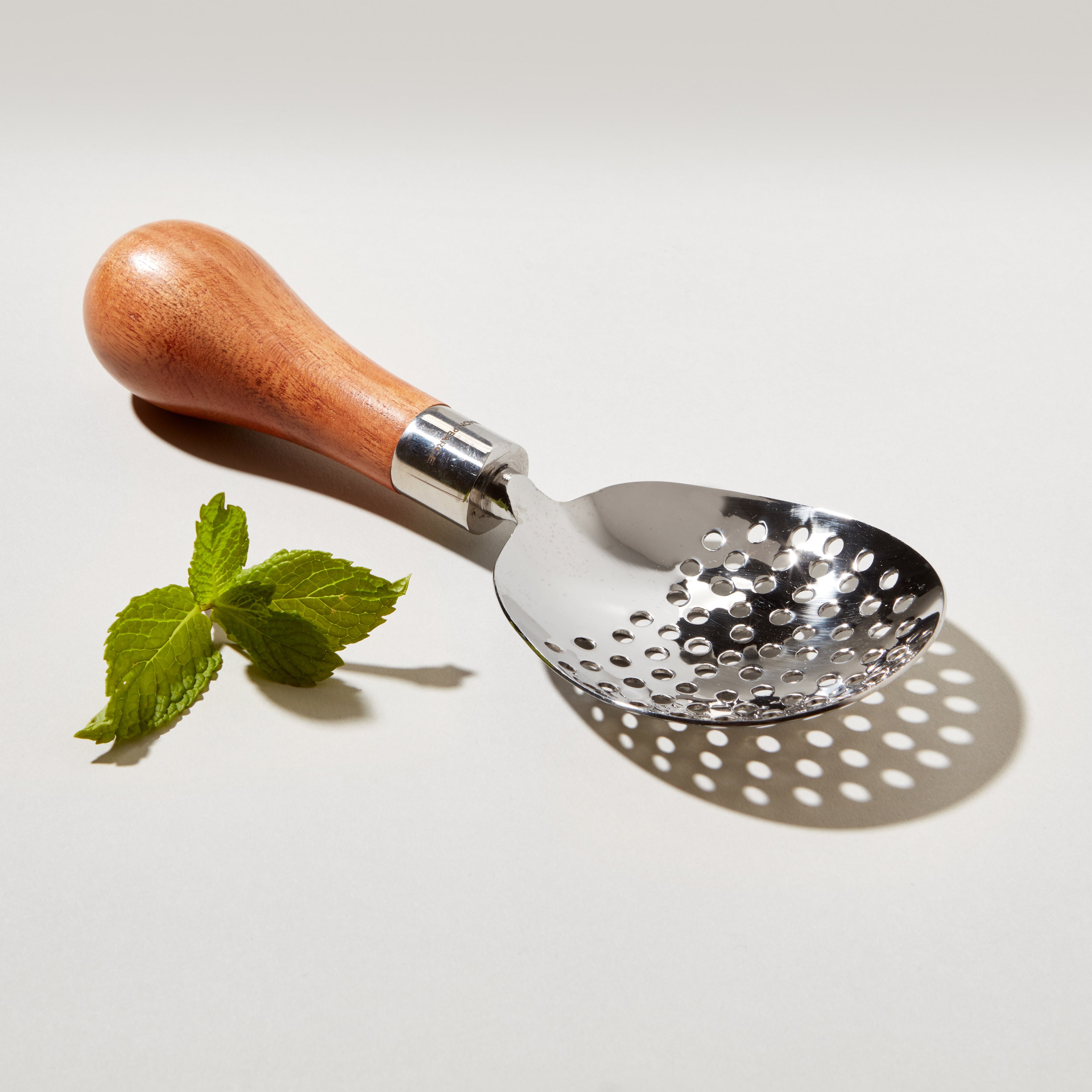 Silver-toned julep strainer with perforated bowl, wooden handle, and metal ferrule, displayed with fresh mint leaves.