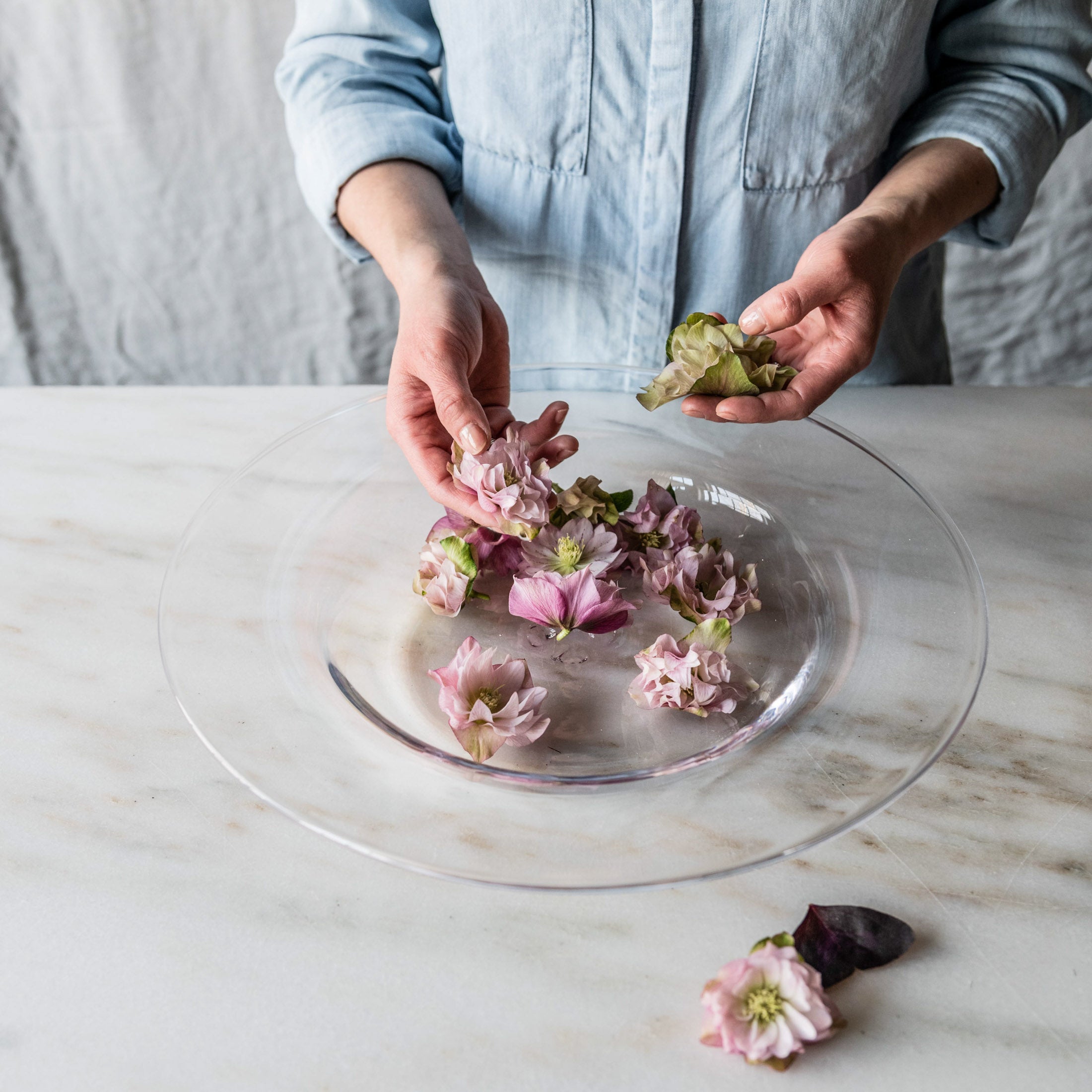 Clear glass platter with person arranging pink and purple edible flowers on marble surface.