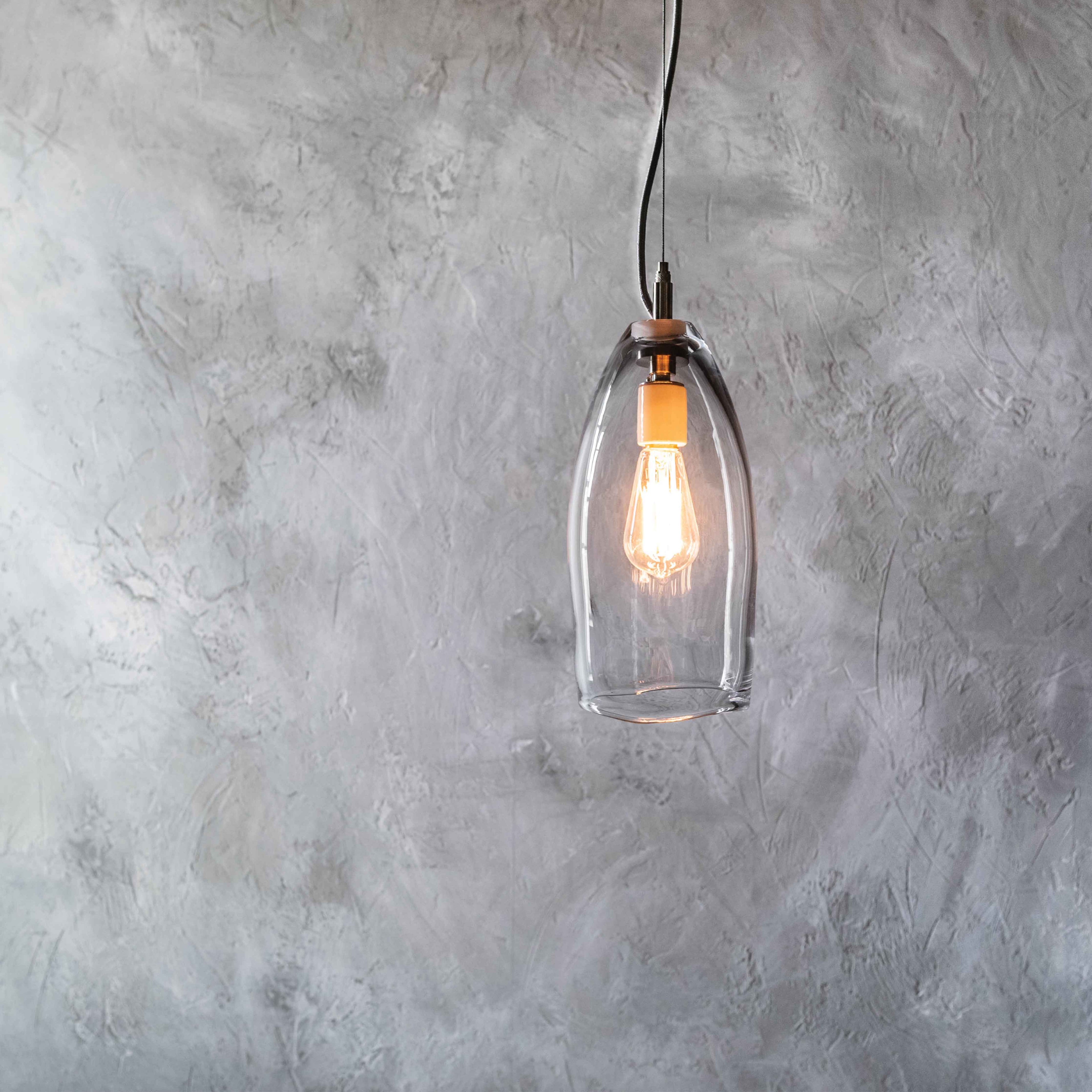 Tall clear glass pendant light with Edison-style bulb and copper fixture hanging from black cord against textured wall.
