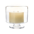 Cream-colored two-wick candle in clear glass hurricane-style holder with pedestal base.