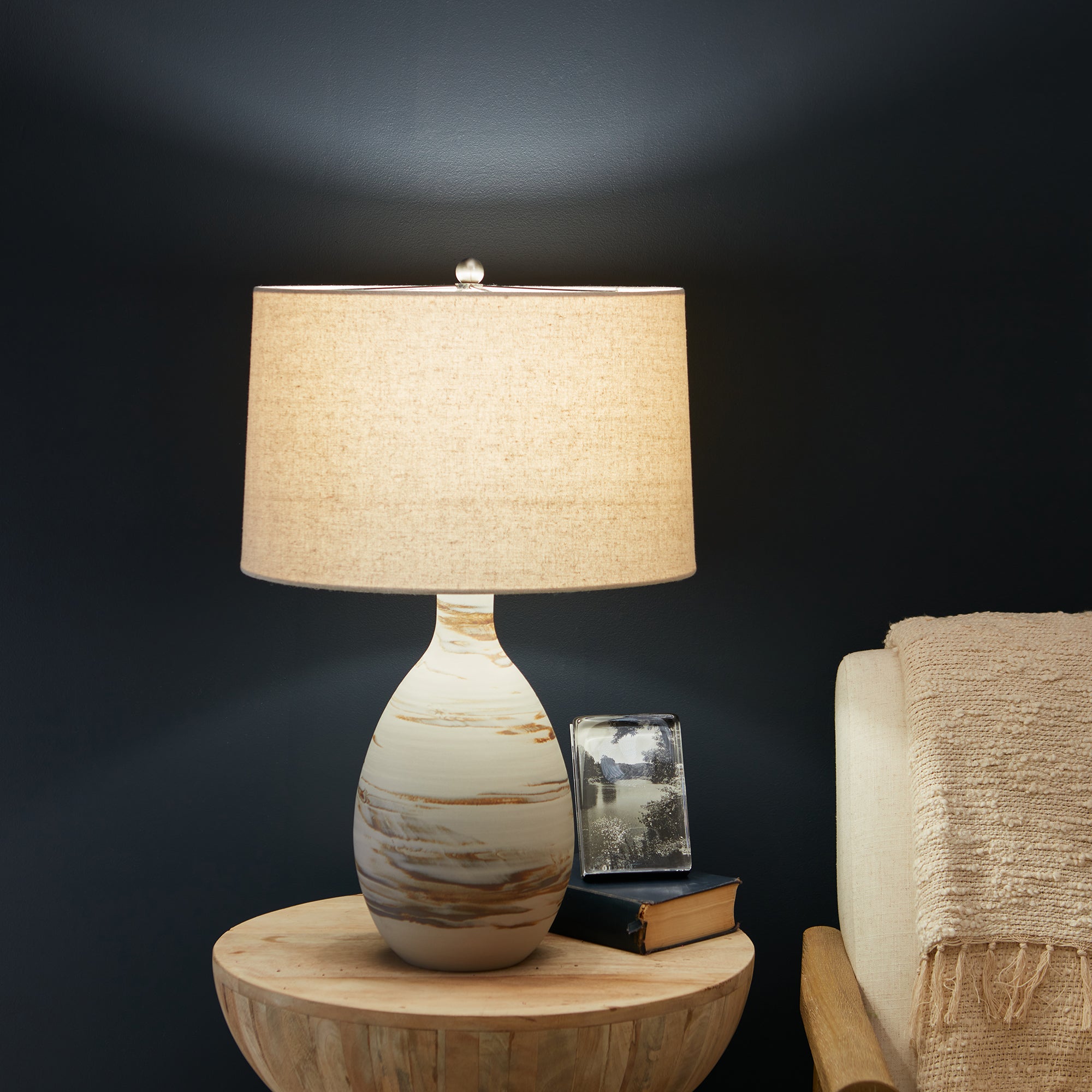 Tall table lamp with cream linen shade and ceramic base featuring brown striations, displayed on wooden table.