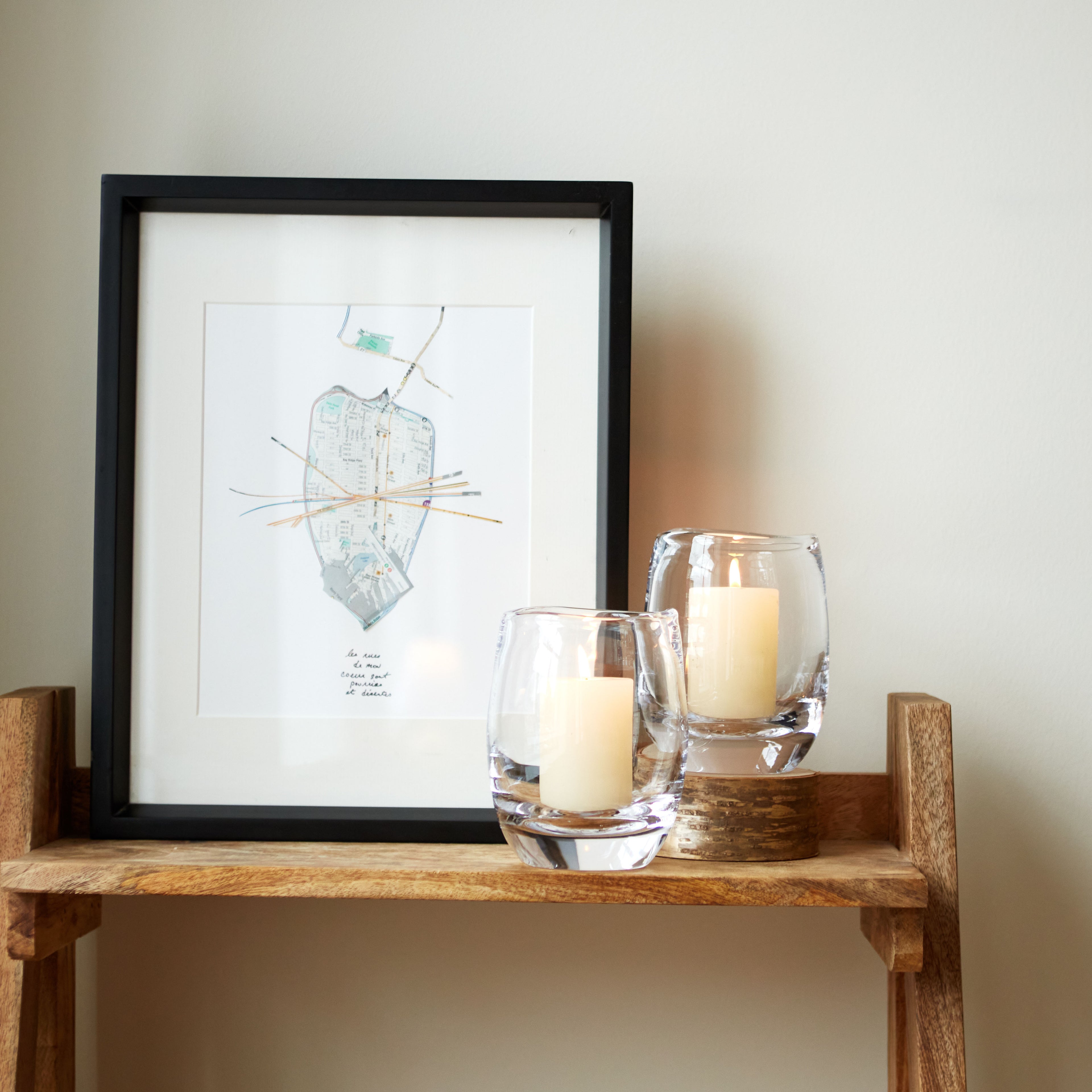 Two clear glass Hurricane candles with cream-colored wax displayed next to a framed map artwork on a wooden shelf.