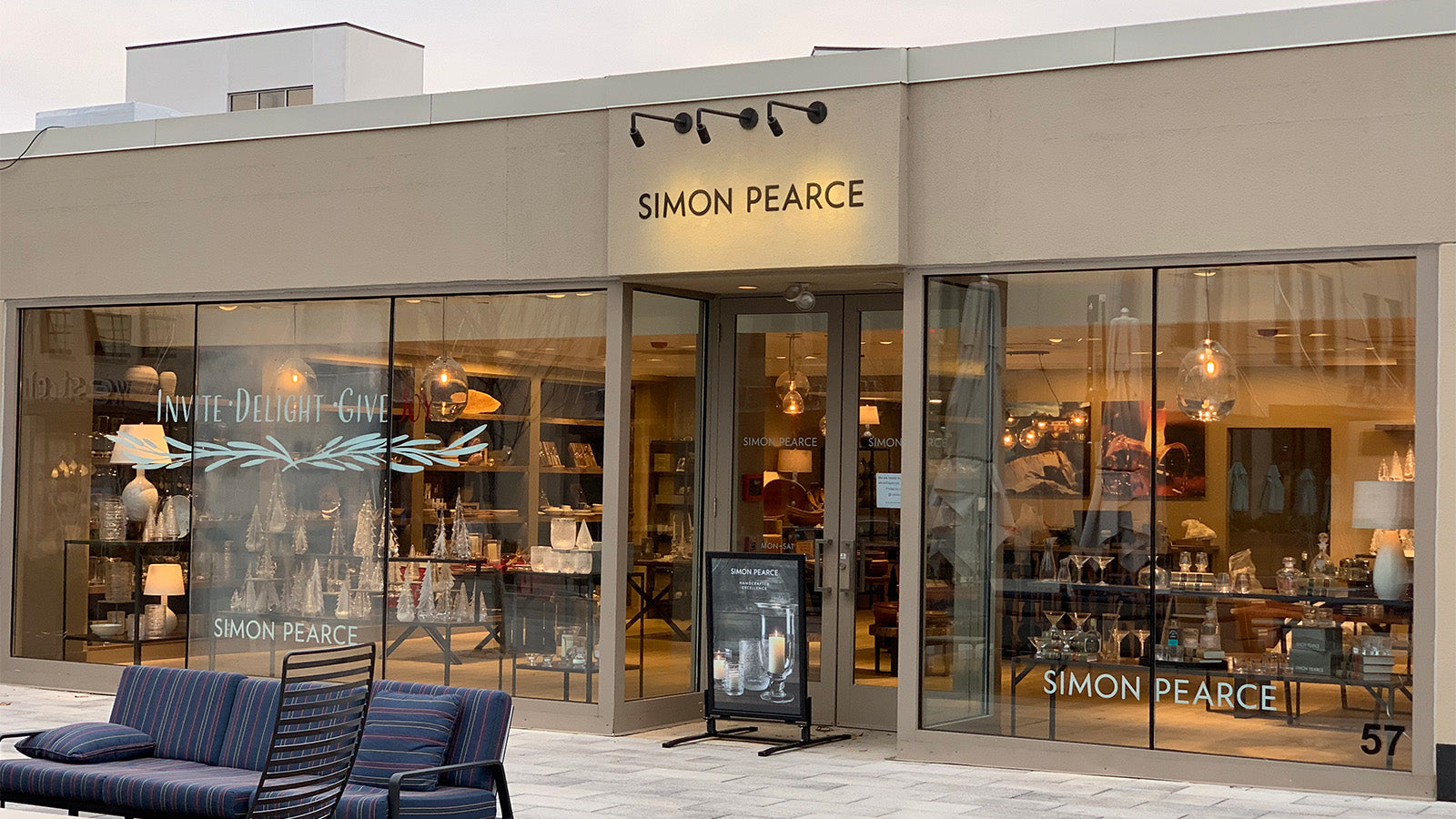 Exterior of the Simon Pearce retail store at 57 St. James Place in Ardmore, Pennsylvania, a modern storefront with large glass windows and seasonal window graphics reading 'Invite, Delight, Give'
