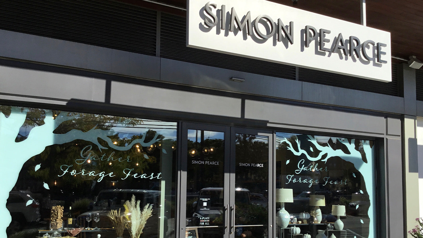 Exterior of the Simon Pearce retail store in Chestnut Hill, Massachusetts, a modern storefront with a backlit sign and seasonal window display reading 'Gather, Forage, Feast'