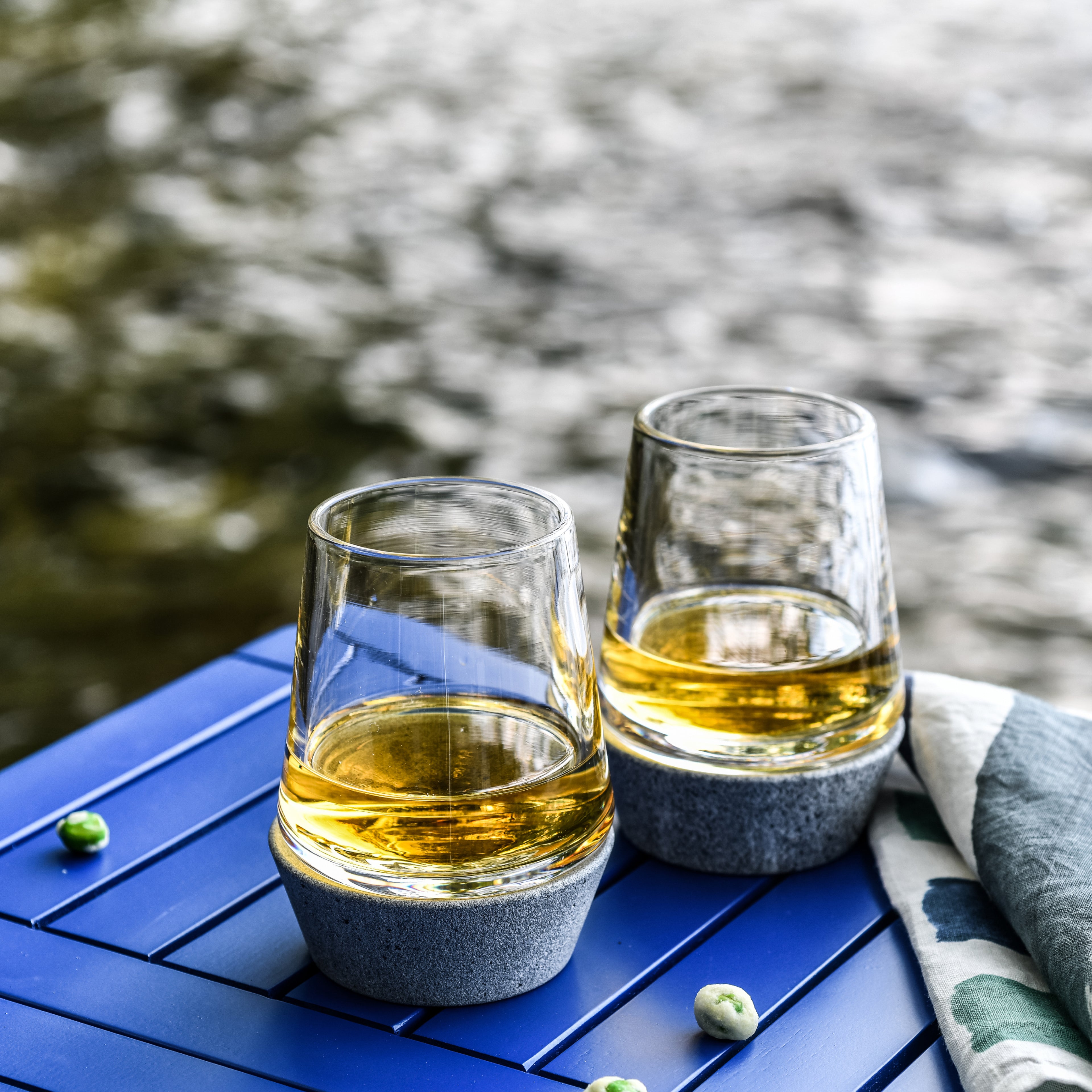 Two glasses filled with golden whiskey sit on a blue surface with gray stone bases and a cloth napkin beside them.