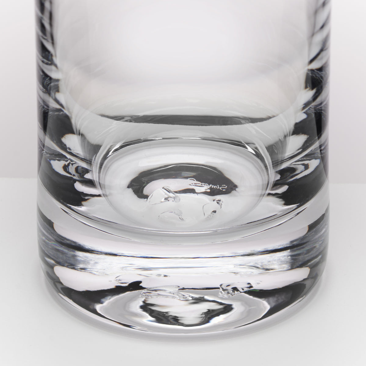 A close-up of a clear glass tumbler with a thick, textured base, showing reflections and the brand name 'Simon Pearce' printed on the glass near the base.